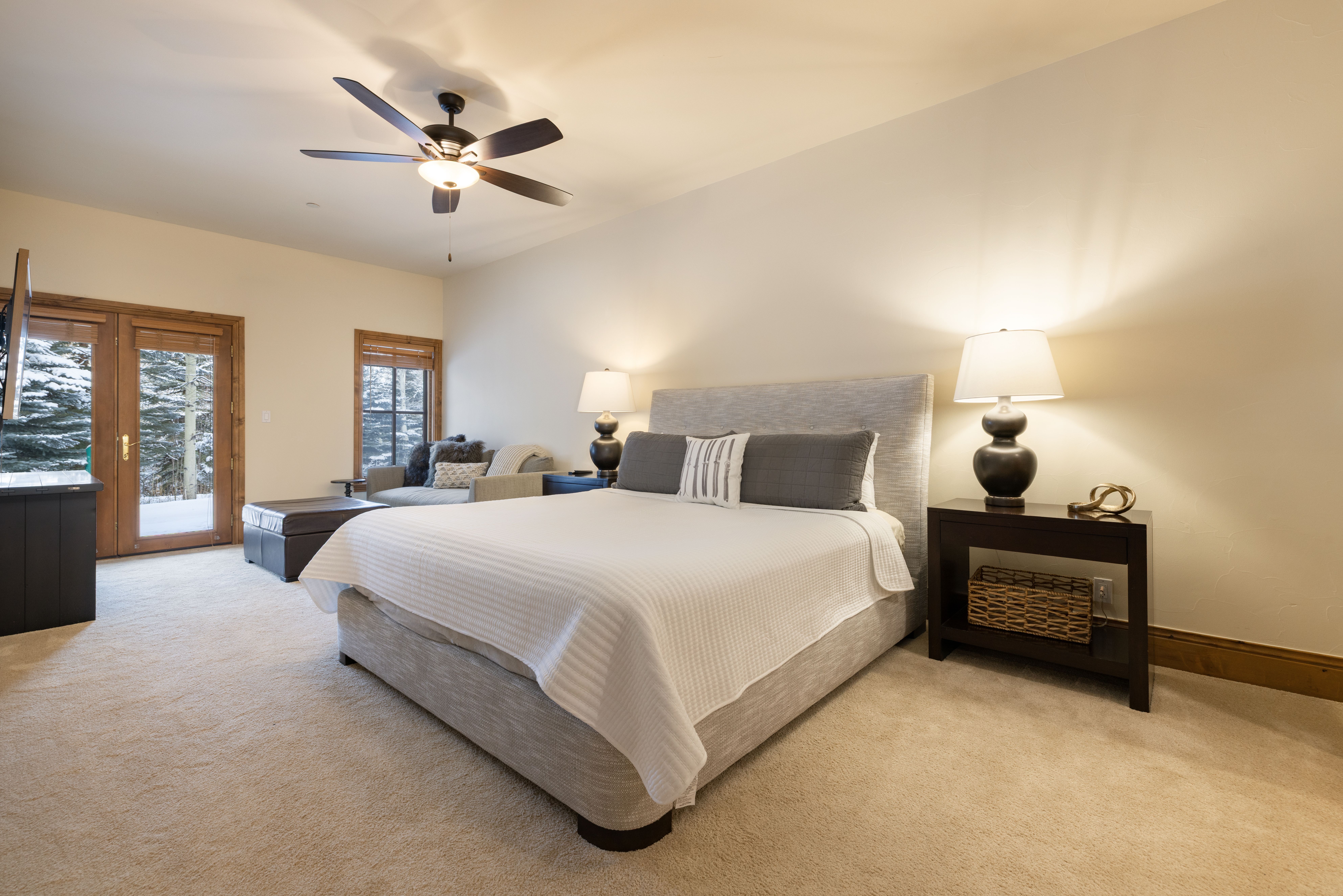 This spacious primary bedroom features a plush, upholstered king-sized bed with a crisp white coverlet and neutral-toned accent pillows. The room is anchored by two dark wood nightstands with matching table lamps, while a cozy seating area with a leather ottoman sits near the glass-paned doors that reveal a snowy outdoor landscape. The neutral color palette, soft carpeting, and warm lighting create a serene and inviting atmosphere.