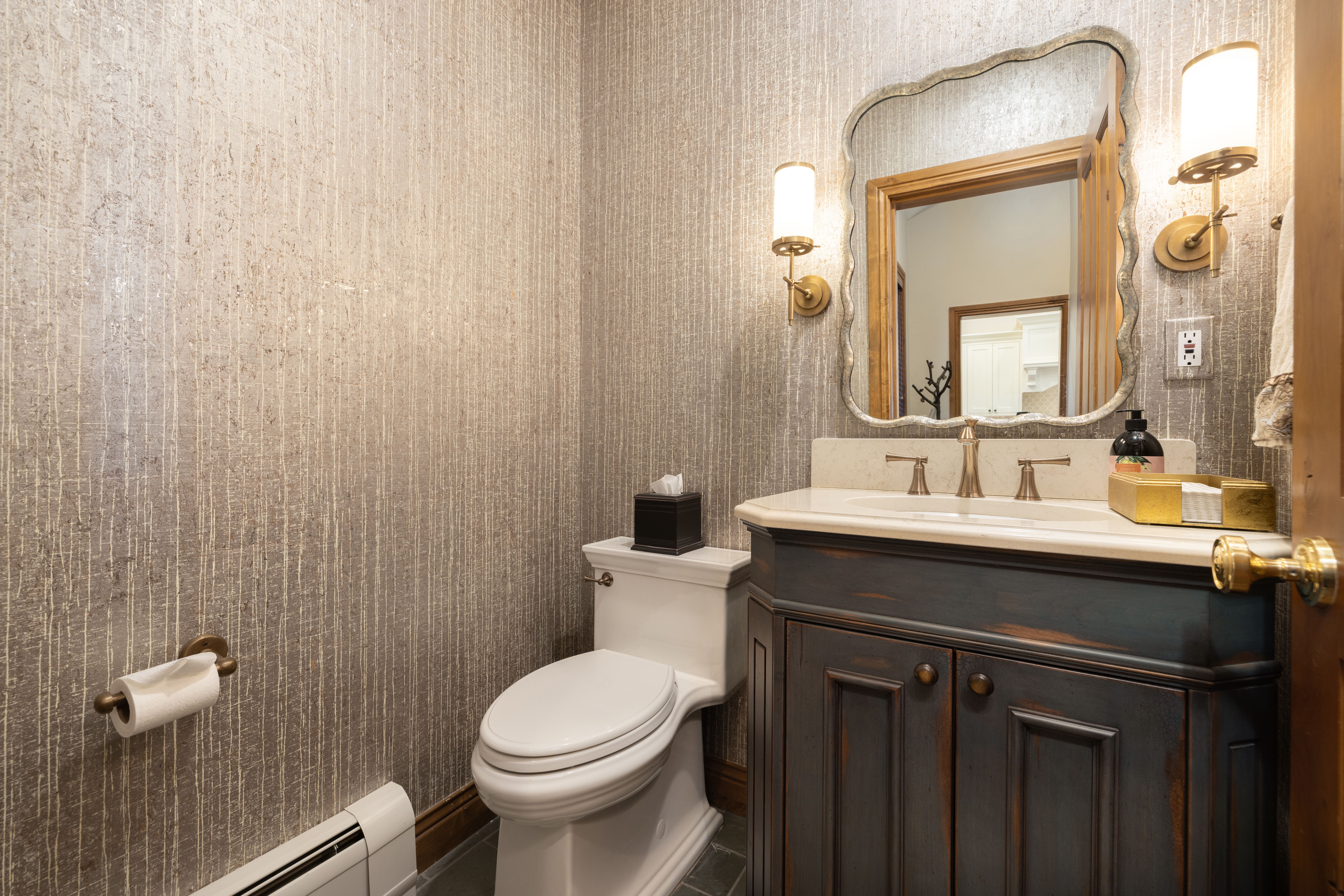 This elegant powder room features textured, metallic-toned wallpaper and a distressed dark wood vanity topped with a light stone countertop. A decorative, scalloped-edge mirror is flanked by two brass wall sconces, creating a sophisticated and warm atmosphere. The space is completed by a white toilet and a classic brass toilet paper holder, offering a refined and compact design.