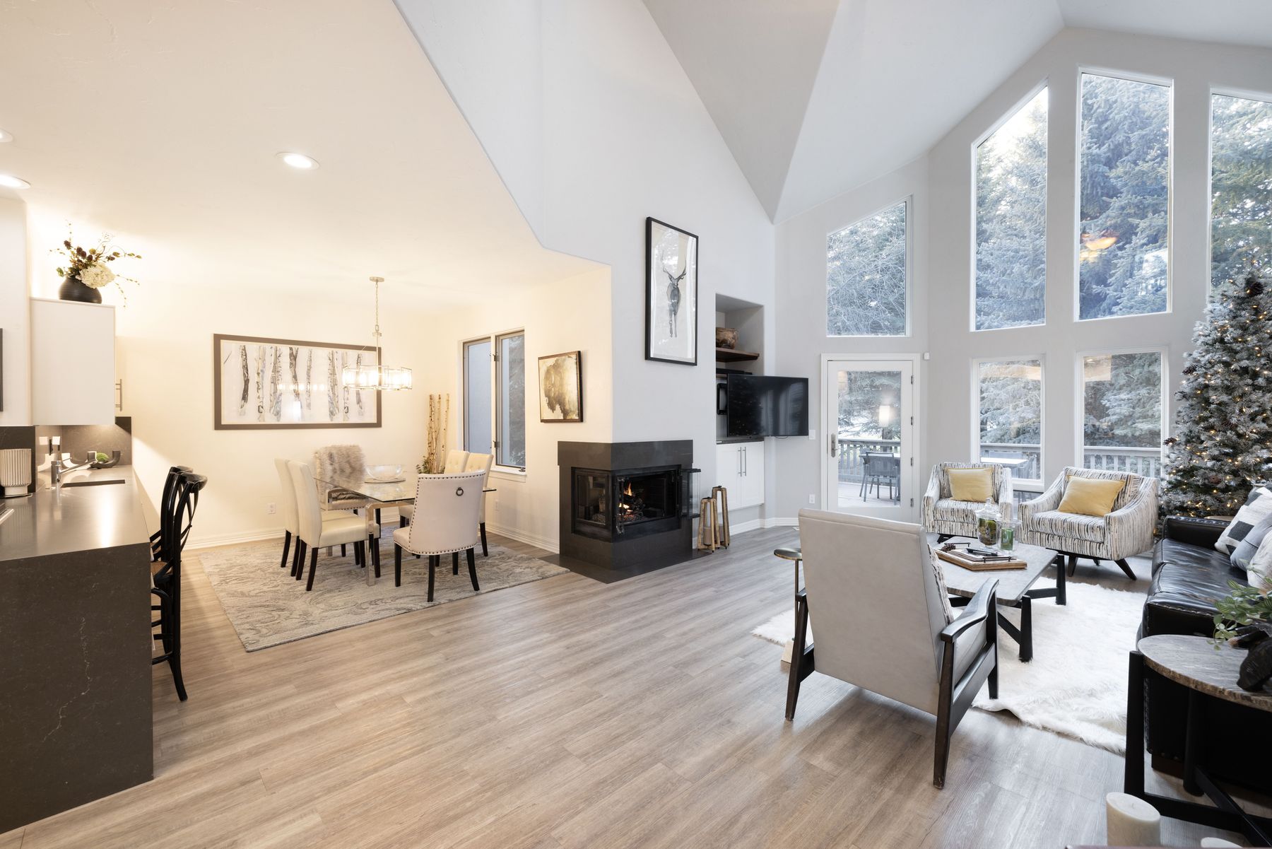 This open-concept living and dining area features vaulted ceilings, light wood-look flooring, and a modern corner fireplace that serves as a central focal point. Large floor-to-ceiling windows flood the space with natural light, showcasing a scenic forest view and a decorated Christmas tree. The room is elegantly furnished with a mix of contemporary seating, a glass-topped dining table, and sophisticated wall art, creating a warm and inviting atmosphere.