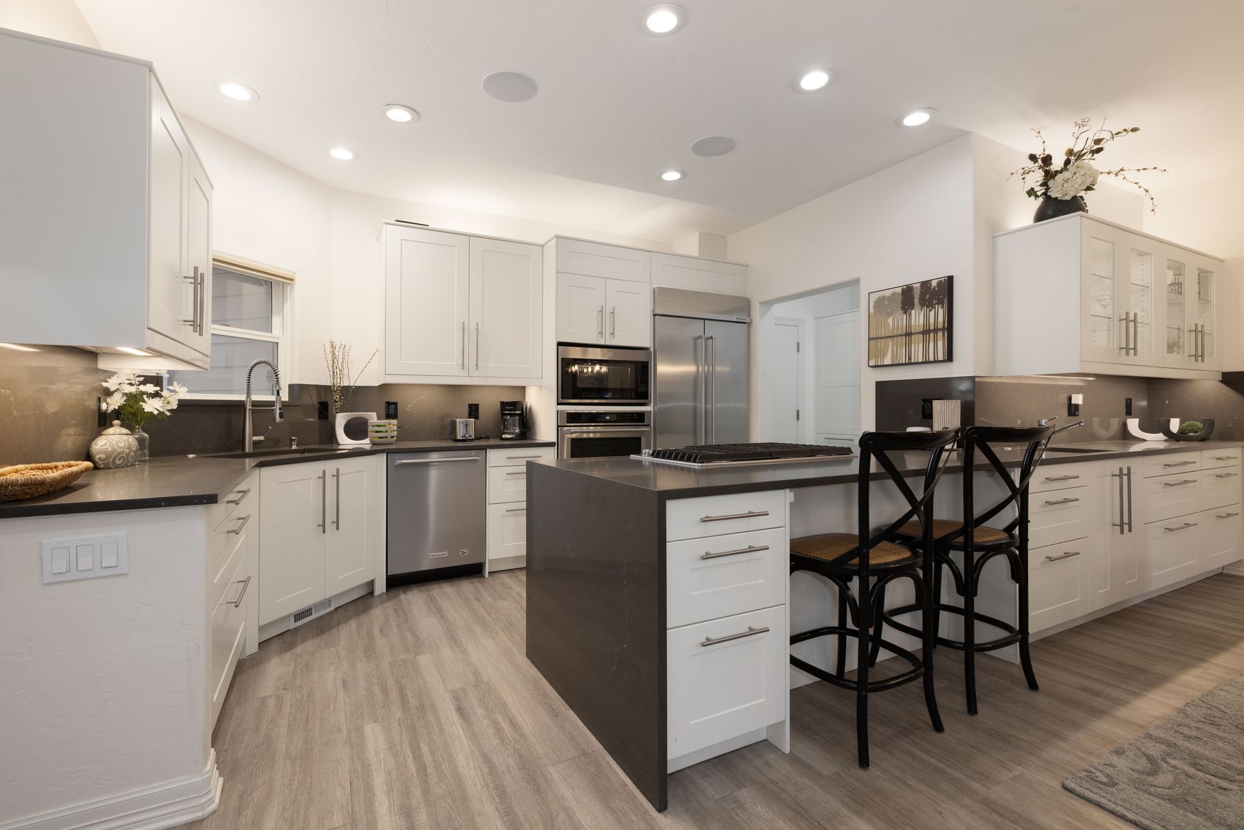 This modern, bright kitchen features crisp white cabinetry contrasted by dark gray quartz countertops and a matching waterfall island. The space is equipped with high-end stainless steel appliances, including a built-in oven and microwave, and is illuminated by recessed ceiling lights. Two black cross-back bar stools provide seating at the island, creating an inviting and functional layout perfect for entertaining.