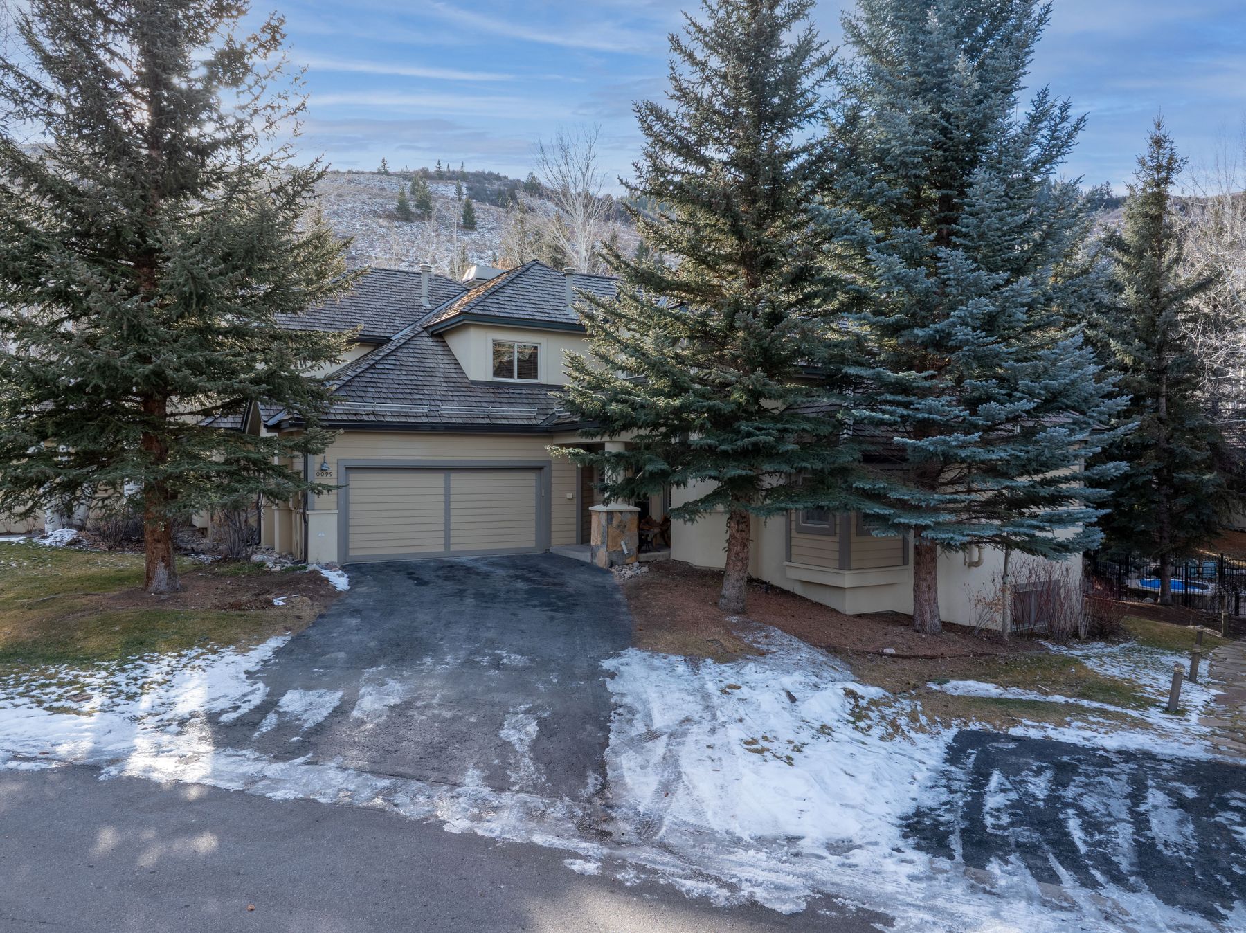 This elevated, eye-level shot captures the front facade of a mountain-style home nestled among tall evergreen trees. The house features a multi-gabled roof, tan siding, and a prominent two-car garage, with a snow-dusted driveway leading up to the entrance. The surrounding landscape, including the hillside in the background, suggests a serene, cold-weather residential setting.