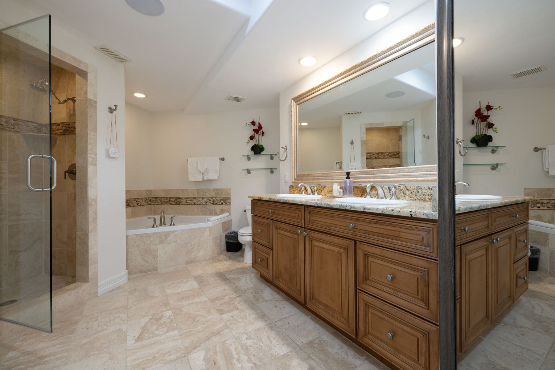 This spacious primary bathroom features a double-sink vanity with warm wood cabinetry and granite countertops, complemented by a large, ornate-framed mirror. The room includes a separate glass-enclosed walk-in shower and a corner soaking tub, all finished with elegant travertine tile flooring and wall accents. The perspective captures the entire layout, highlighting a clean, luxurious, and well-lit atmosphere.
