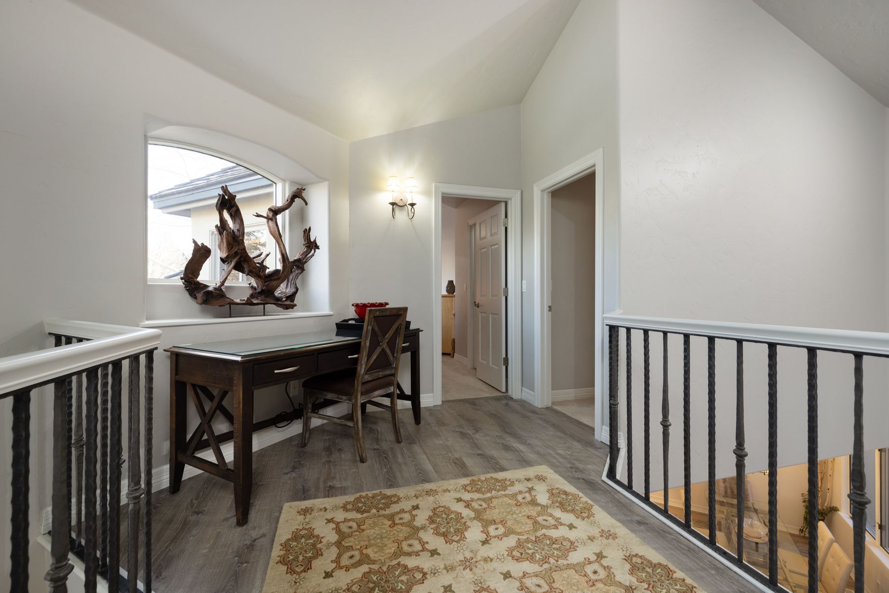 This cozy home office nook is situated on an upper-level landing, featuring a dark wood desk paired with a matching cross-back chair. A unique piece of driftwood art sits on the windowsill, while a patterned area rug adds warmth to the wood-look flooring. The space is framed by wrought-iron railings and leads into adjacent rooms, creating an inviting and functional transition area.