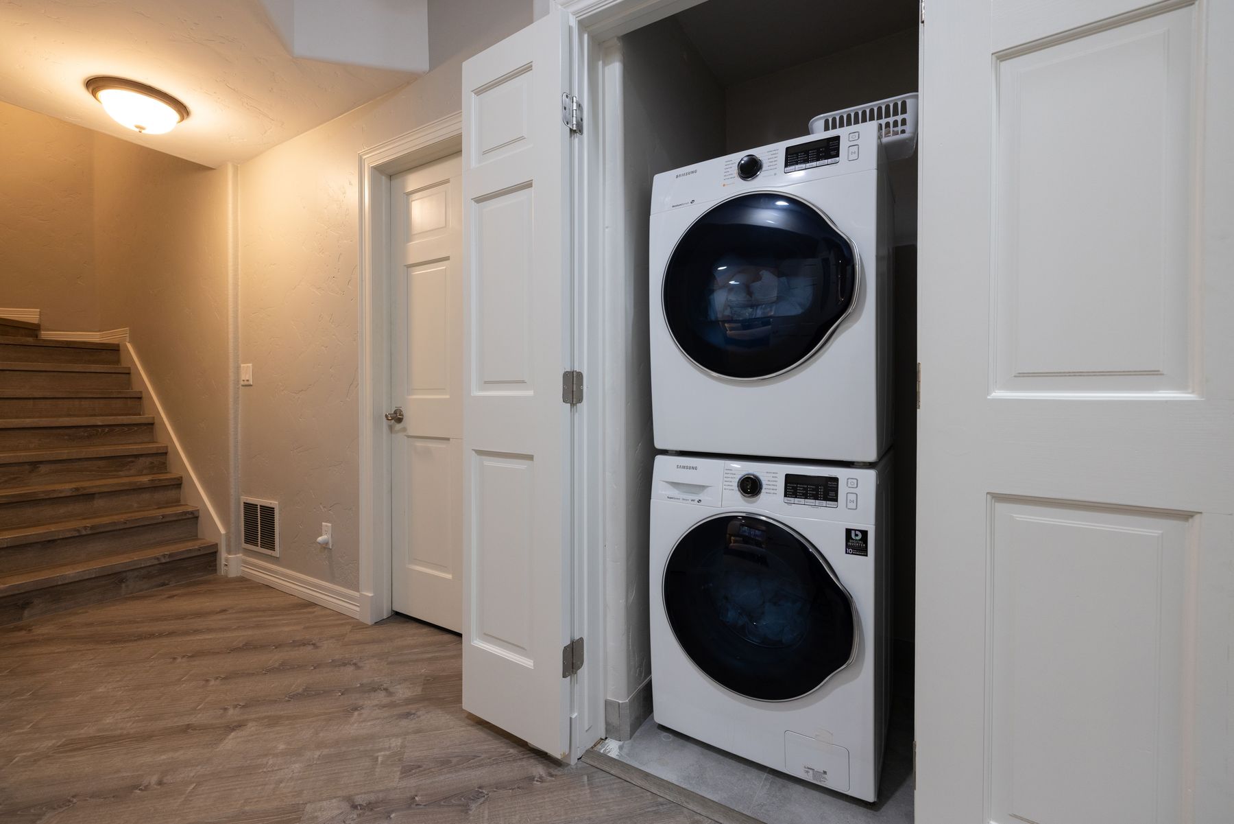 This image captures a compact laundry area tucked into a closet space, featuring a stacked Samsung washer and dryer unit. To the left, a set of wooden stairs leads upward, and a closed white door is visible, all set against a neutral-toned hallway with wood-look flooring. The perspective is eye-level, highlighting the efficient use of space in a modern residential setting.