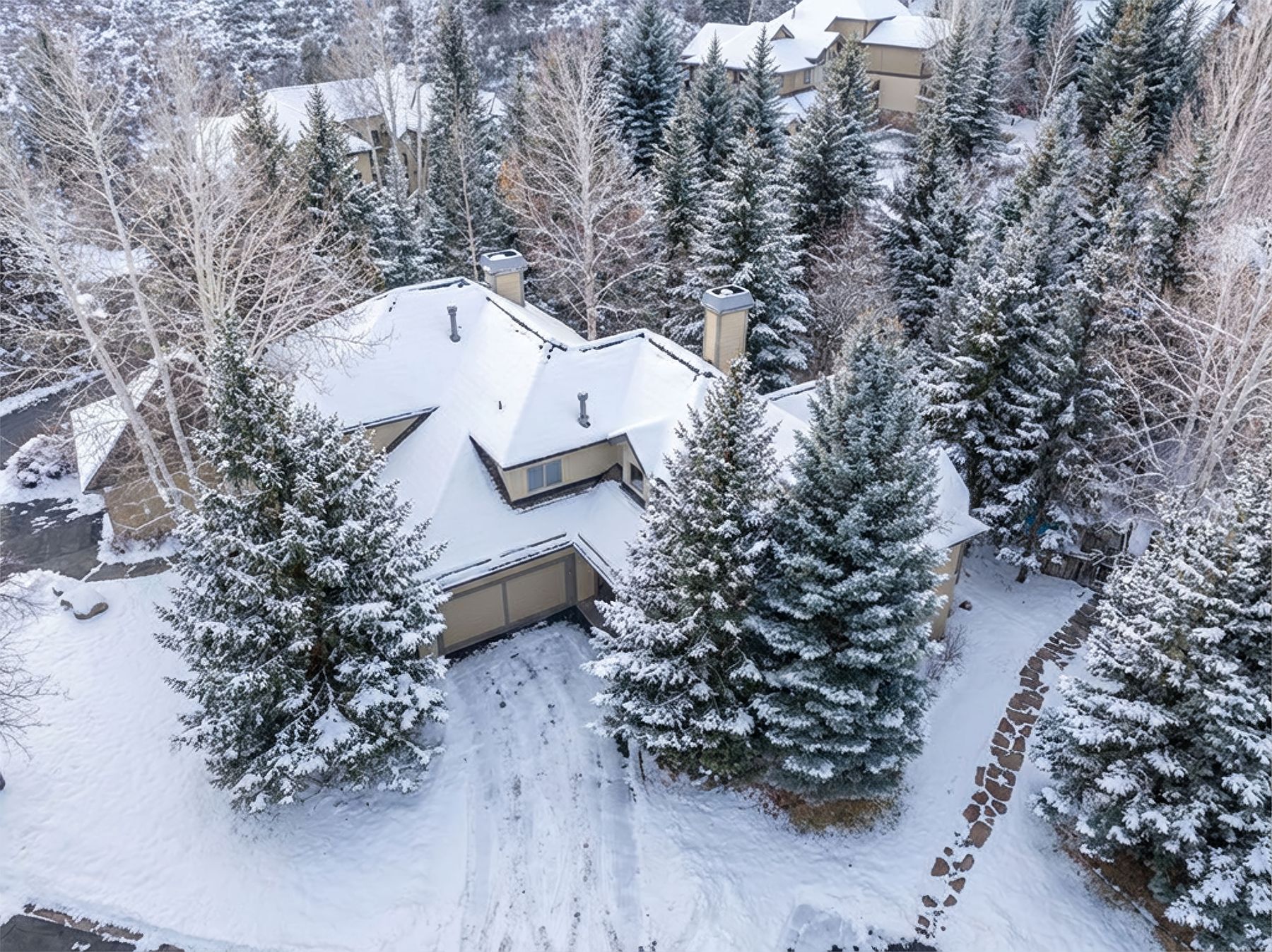 This high-angle aerial shot captures a charming mountain home nestled within a dense, snow-covered forest of evergreen and deciduous trees. The house features a multi-gabled roof blanketed in fresh snow, a two-car garage, and a stone-paved walkway leading through the wintry landscape. The scene evokes a serene, secluded atmosphere, highlighting the property's integration into a picturesque, cold-weather environment.