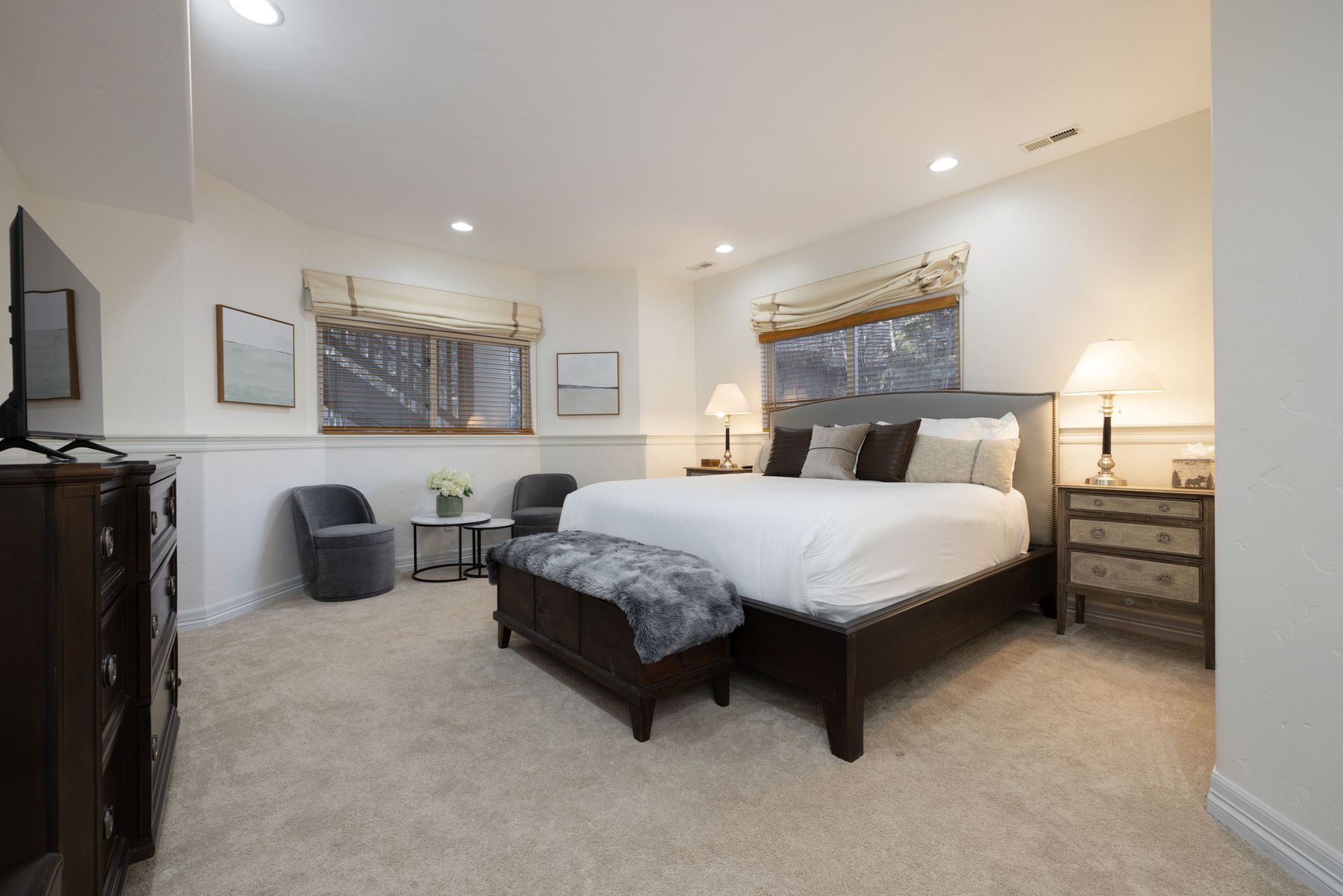 This spacious primary bedroom features a large dark wood bed frame with a plush white duvet and a grey faux-fur throw, complemented by matching dark wood nightstands and a dresser. The room is accented by two grey armchairs near a small bistro table, creating a cozy seating area, while neutral walls and soft carpeting provide a calm, inviting atmosphere. The perspective is a wide-angle shot from the doorway, showcasing the balanced layout and elegant, traditional decor.