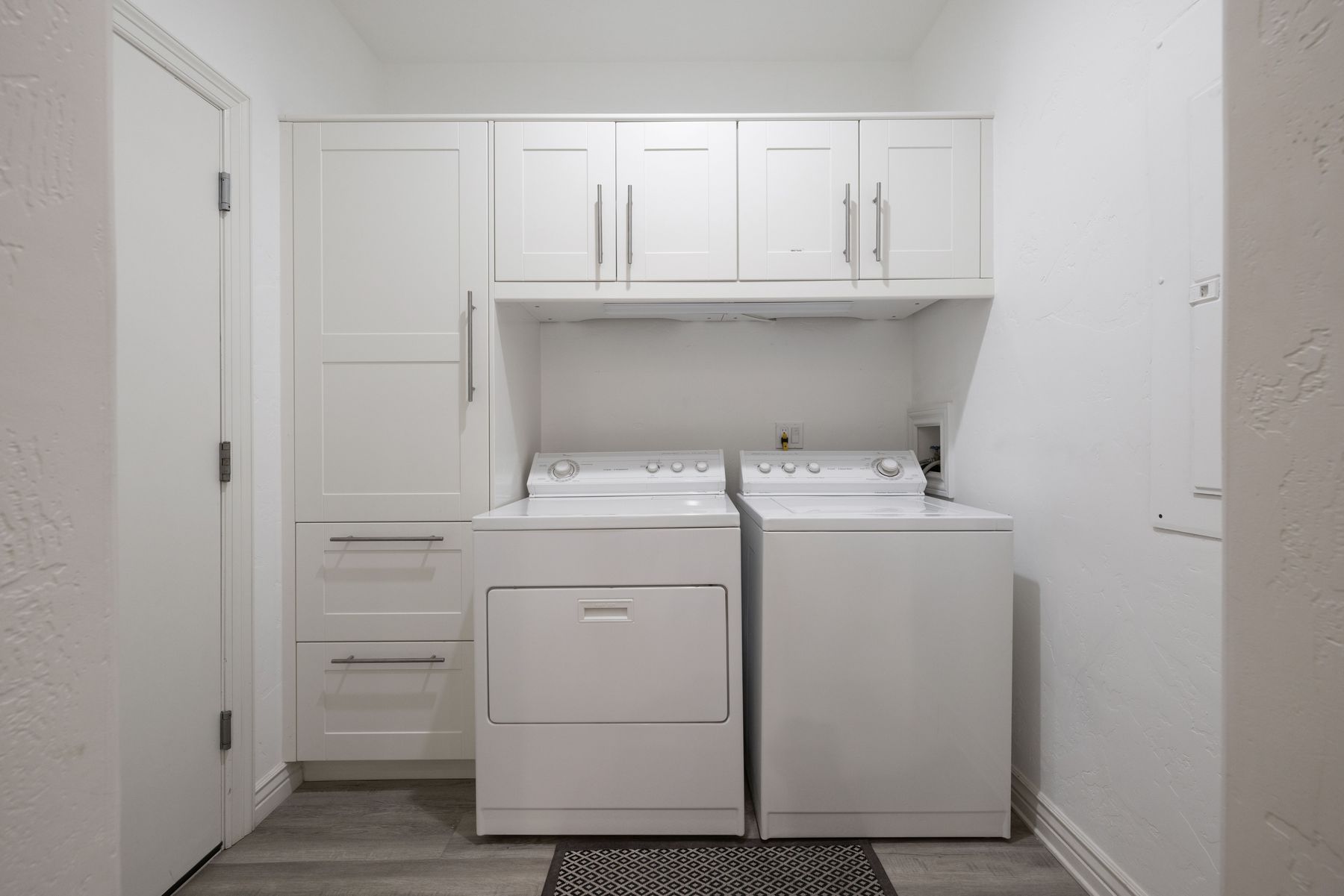 This clean and functional laundry room features white cabinetry with ample storage, including a tall pantry-style cabinet and two lower drawers. A side-by-side washer and dryer set sits neatly beneath wall-mounted cabinets, all set against a neutral white backdrop with light wood-look flooring. The space is well-organized and bright, offering a practical and modern aesthetic for a home utility area.