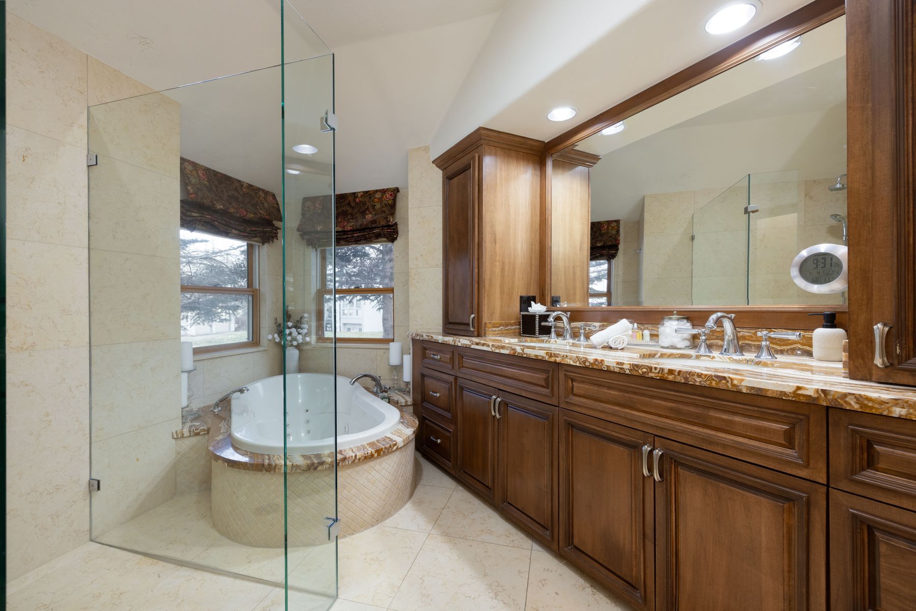 This luxurious primary bathroom features a warm, traditional aesthetic with rich wood cabinetry and a striking, multi-toned marble countertop. A deep soaking jetted tub is nestled into a tiled alcove next to a glass-enclosed shower, creating a spa-like atmosphere. The space is well-lit by recessed lighting and a large mirror, emphasizing the elegant tile flooring and high-end finishes.