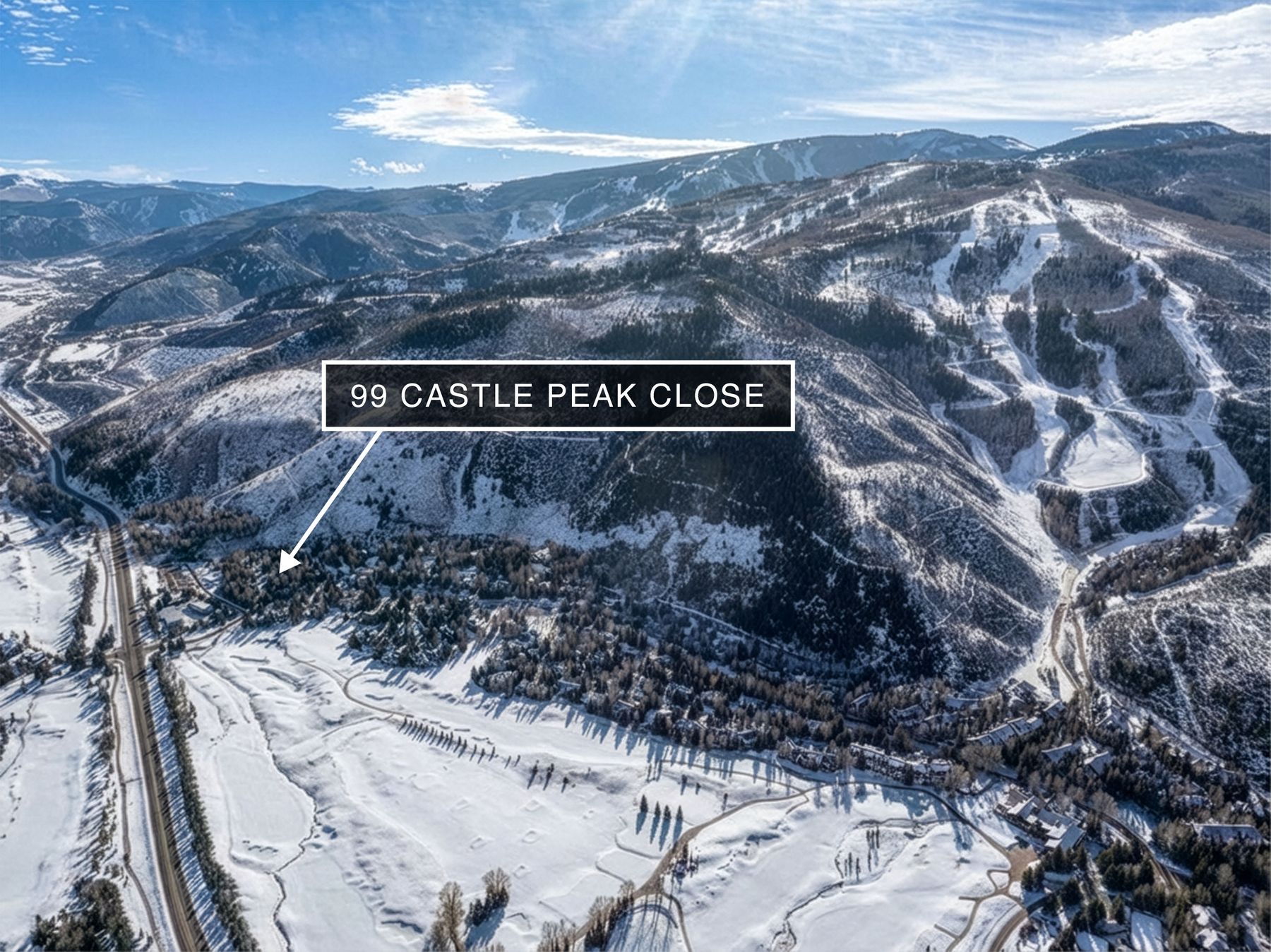 This high-altitude aerial shot captures a sprawling, snow-covered mountain landscape, highlighting the location of 99 Castle Peak Close nestled near a ski resort. The perspective emphasizes the vastness of the terrain, featuring dense evergreen forests, winding roads, and the proximity to expansive ski slopes. The overall impression is one of a premier, secluded alpine retreat set within a dramatic winter wonderland.
