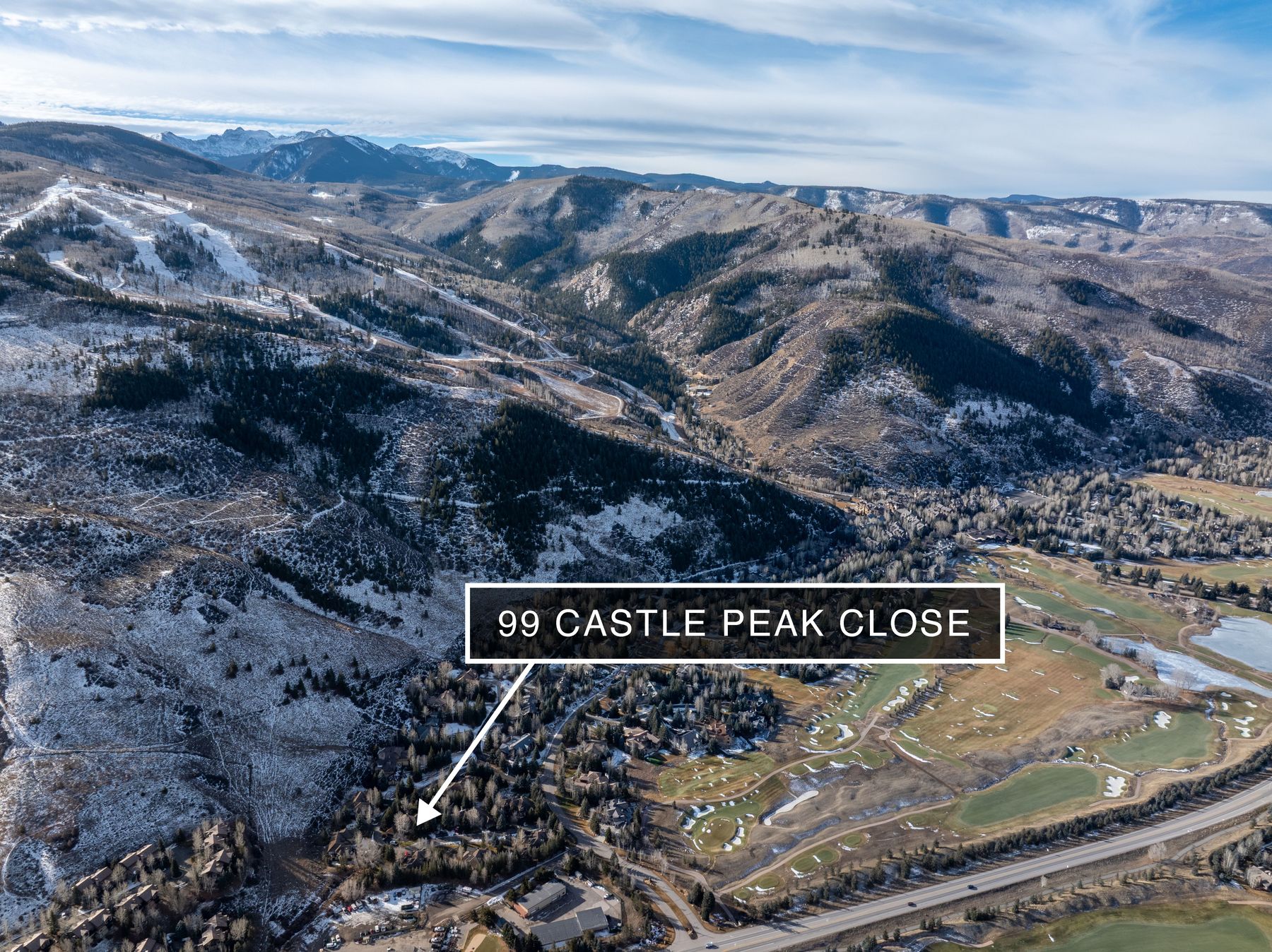 This high-altitude aerial shot captures a sprawling mountain landscape, highlighting the property's location at 99 Castle Peak Close nestled among dense trees. The scene showcases a vast valley with a golf course, winding roads, and dramatic, snow-dusted mountain peaks in the background. The perspective provides a grand sense of scale, emphasizing the serene, secluded, and scenic nature of the residential setting.