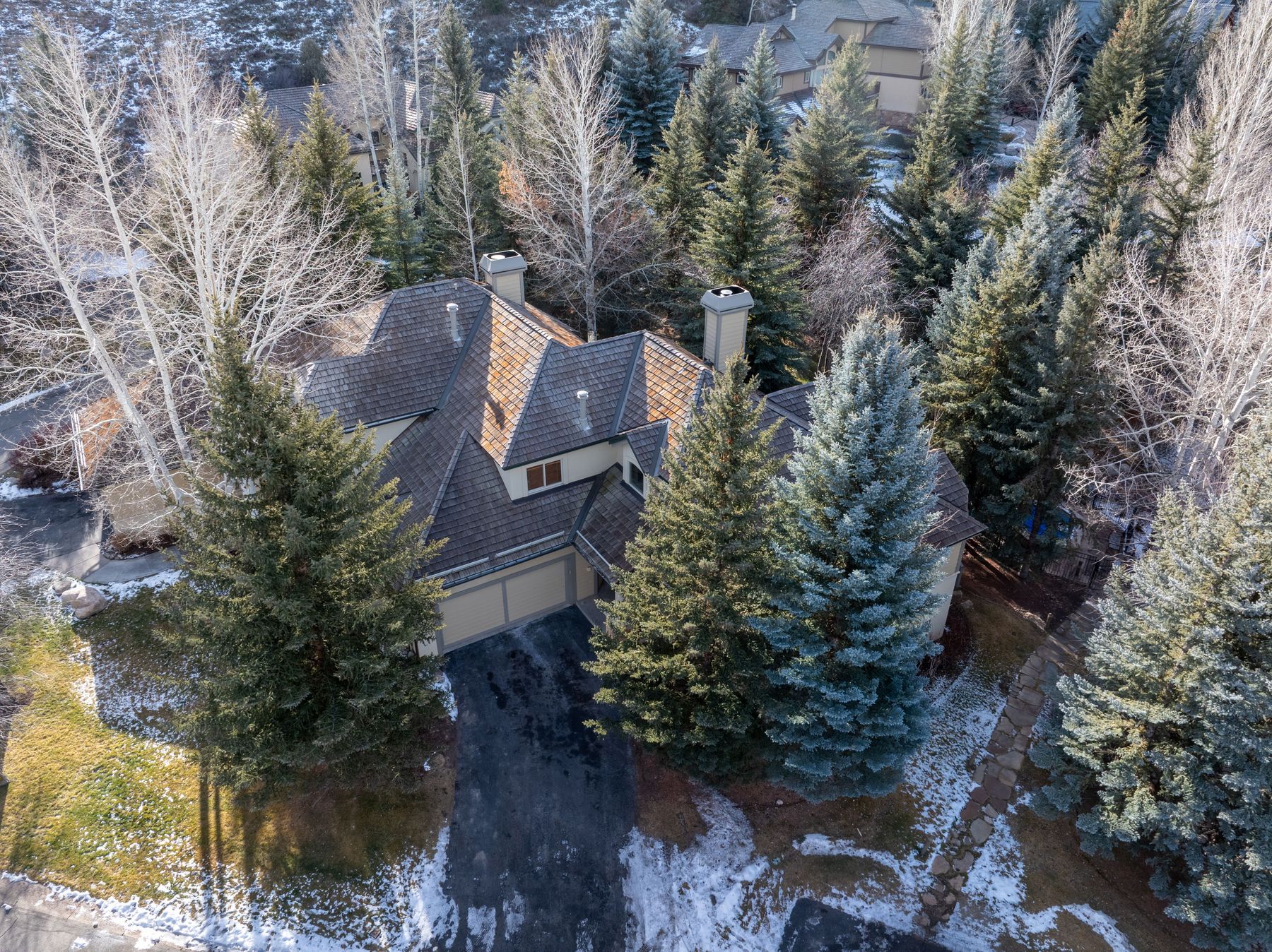 This high-angle aerial perspective captures a multi-story mountain-style home nestled within a dense, evergreen-rich landscape. The residence features a complex, multi-gabled roofline with dark shingles, a two-car garage, and a paved driveway, all surrounded by a mix of mature pine trees and bare-branched aspens. The scene conveys a serene, secluded atmosphere, characteristic of a high-end mountain retreat during the late winter or early spring season.
