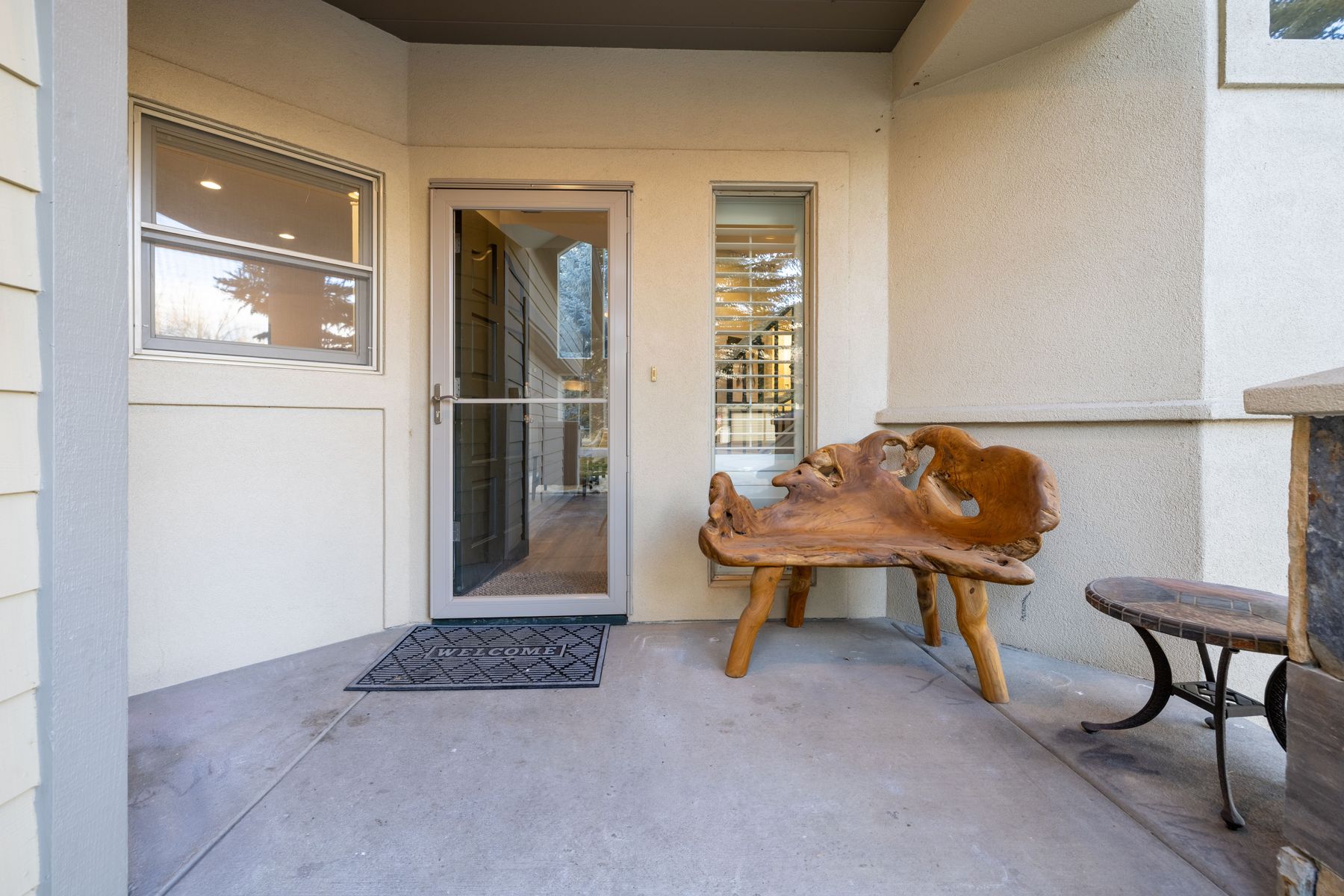 This inviting front entryway features a concrete porch adorned with a unique, organic-shaped wooden bench and a small side table. A glass storm door leads into the home, flanked by a large window and a narrow, shuttered window, creating a bright and welcoming atmosphere. The neutral stucco exterior and clean lines provide a warm, well-maintained first impression for potential buyers.