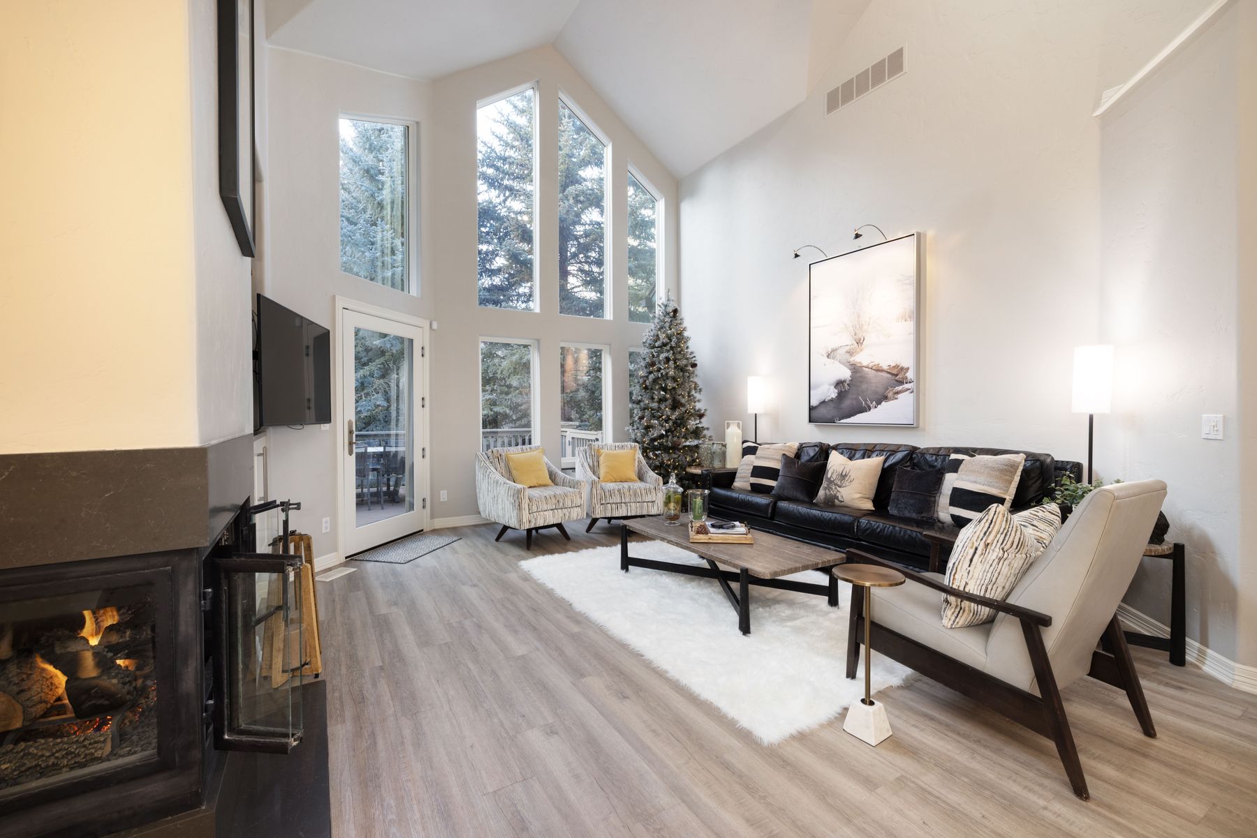 This spacious, high-ceilinged living room features a modern aesthetic with a large black leather sofa, a cozy white faux-fur rug, and a rustic wooden coffee table. The room is anchored by a striking floor-to-ceiling fireplace on the left and expansive windows that flood the space with natural light, showcasing a serene forest view. The neutral color palette and thoughtful decor, including a large landscape art piece, create an inviting and sophisticated atmosphere.