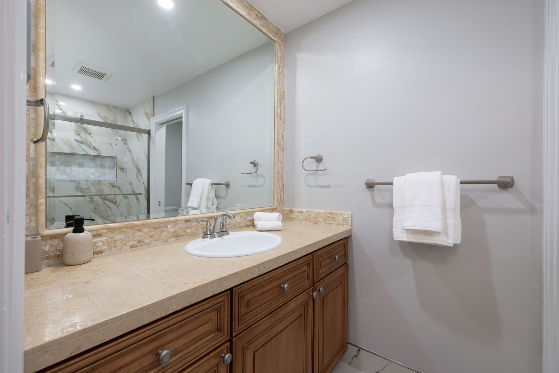 This bathroom features a warm, traditional aesthetic with a wooden vanity topped by a beige stone countertop and an integrated sink. A large, framed mirror reflects the shower area, which is finished with elegant marble-patterned tiles, while a towel bar with a clean white towel adds a functional touch to the neutral-toned wall. The space is well-lit and presents a clean, inviting atmosphere suitable for a guest or secondary bathroom.