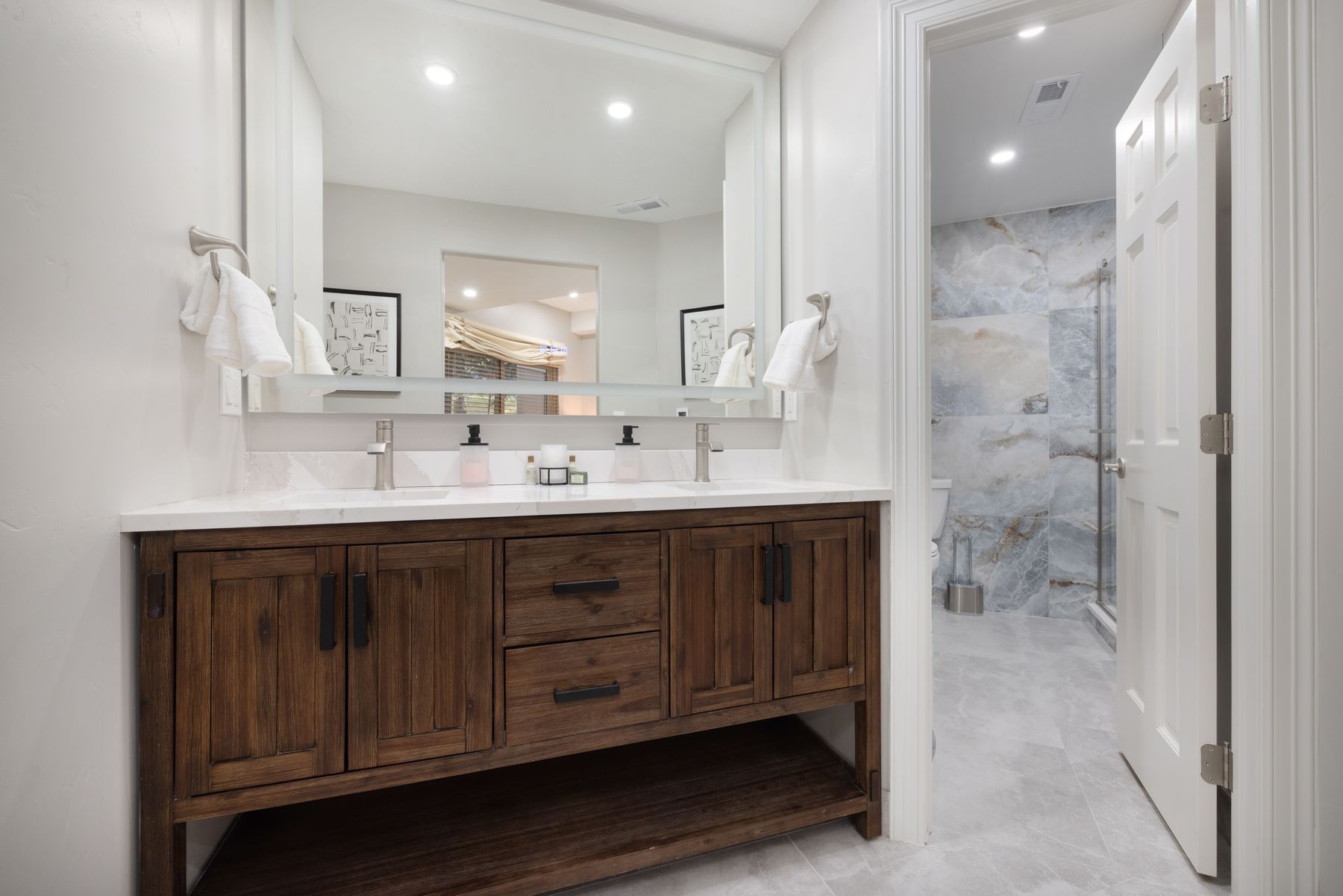 This primary bathroom features a rustic-style wooden double vanity with a clean white countertop and a large, modern mirror. The space is bright and well-lit, with a doorway leading into a separate area containing a shower with striking blue-veined marble tile work. The overall aesthetic blends warm, natural wood tones with contemporary fixtures for a sophisticated and inviting atmosphere.