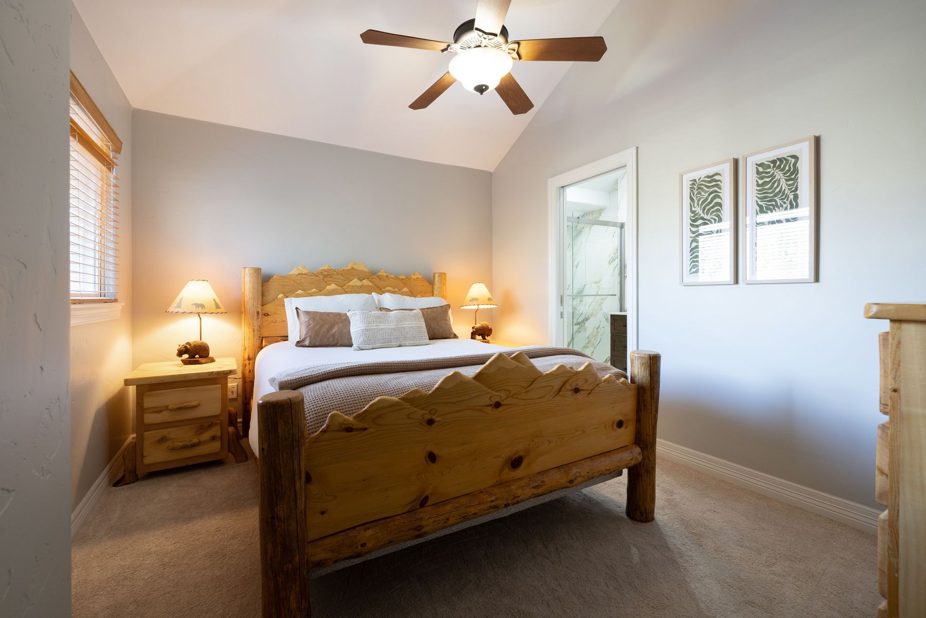 This cozy bedroom features a rustic log-style bed frame with a mountain-inspired headboard and footboard, creating a warm, cabin-like atmosphere. The room is accented by matching wooden nightstands with bear-themed lamps, neutral bedding, and two framed botanical prints on the wall. A ceiling fan provides overhead lighting, and a doorway leads into an adjacent bathroom, enhancing the functional layout of this inviting space.