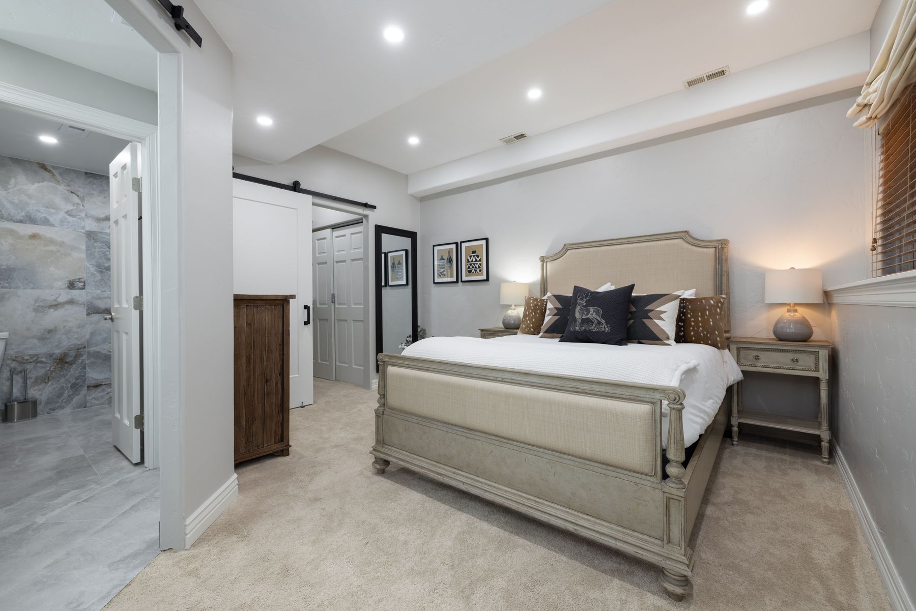This primary bedroom features a sophisticated, neutral color palette with a large upholstered bed as the focal point, flanked by matching nightstands and lamps. The room includes a seamless transition into an attached bathroom with marble-look tile, and a sliding barn door adds a touch of rustic charm. The space is well-lit with recessed lighting, creating a warm and inviting atmosphere.