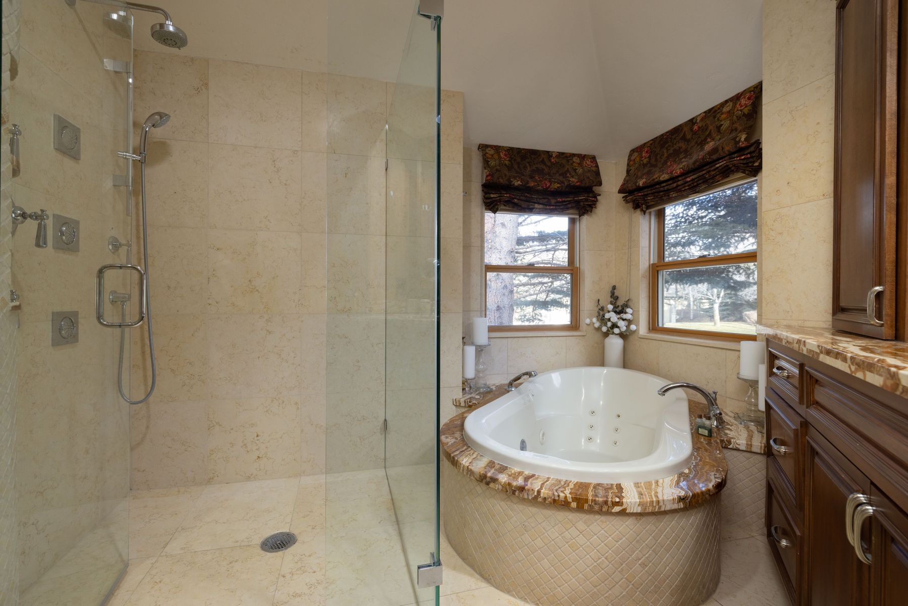 This luxurious primary bathroom features a spacious walk-in glass shower with multiple showerheads and a separate jetted soaking tub set into a custom stone-tiled platform. The room is finished with elegant beige stone tiling, rich wood cabinetry with marble countertops, and large windows adorned with patterned valances that bring in natural light. The overall aesthetic is sophisticated and spa-like, offering a serene retreat within the home.