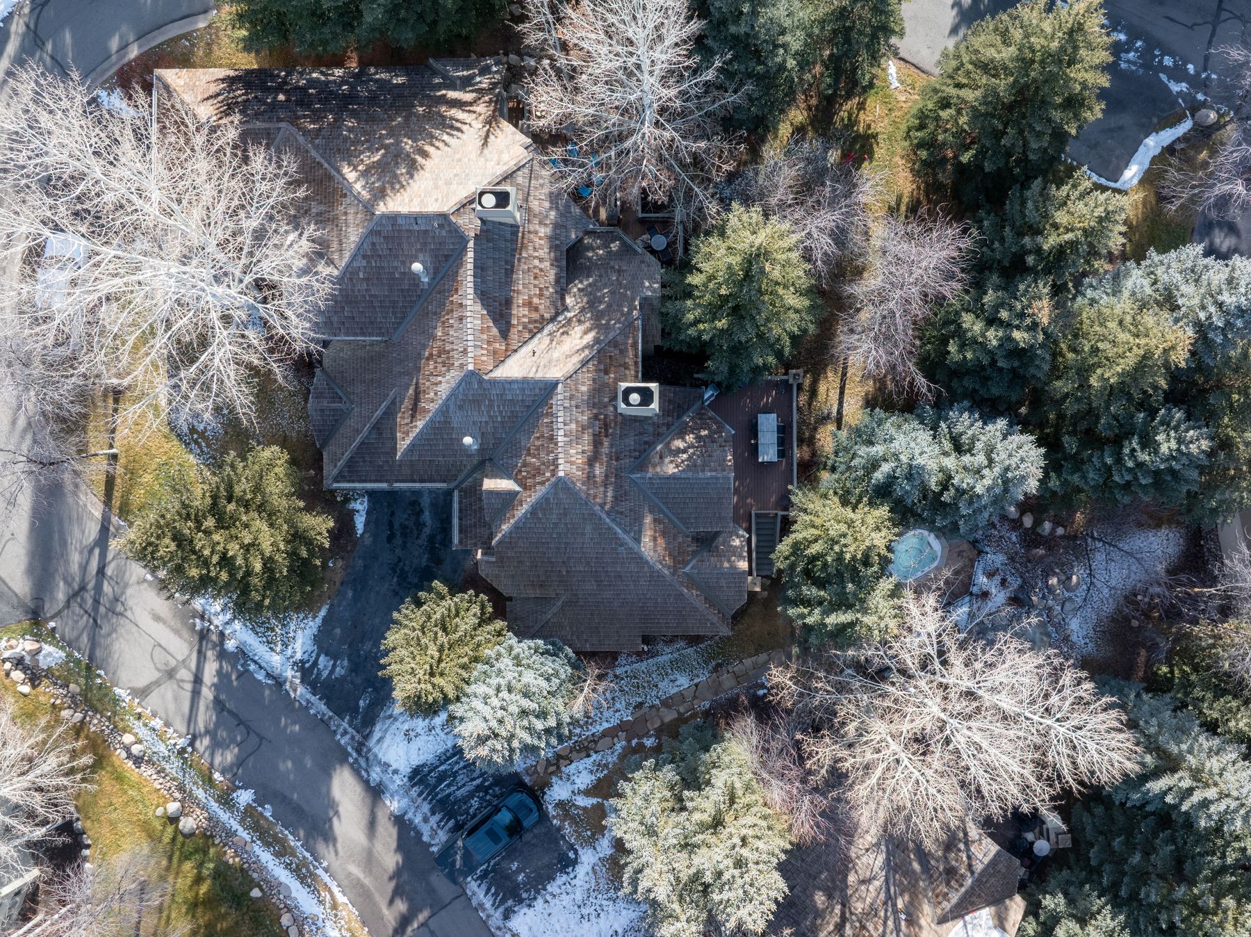 This high-angle aerial view captures a sprawling, multi-gabled residential home nestled within a wooded, snow-dusted landscape. The property features a complex shingled roofline, a private driveway with a parked vehicle, and a secluded rear deck area complete with a small spa. The surrounding mature trees and winding road suggest a quiet, established neighborhood setting.