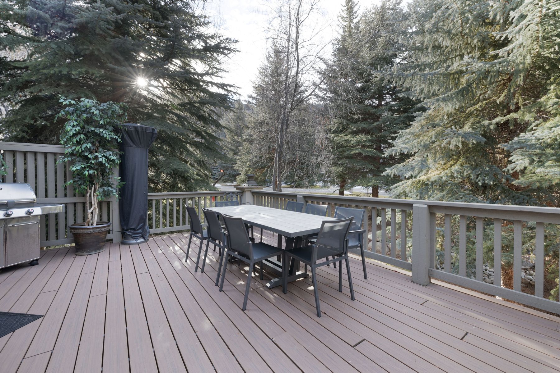 This spacious outdoor deck features composite wood-look decking and a dining set with a rectangular table and six modern chairs. A stainless steel grill and a covered patio heater are positioned to the side, while the deck is enclosed by a sturdy railing overlooking a dense, wooded forest. The scene is captured from a slightly elevated perspective, emphasizing the serene and private atmosphere perfect for outdoor entertaining.