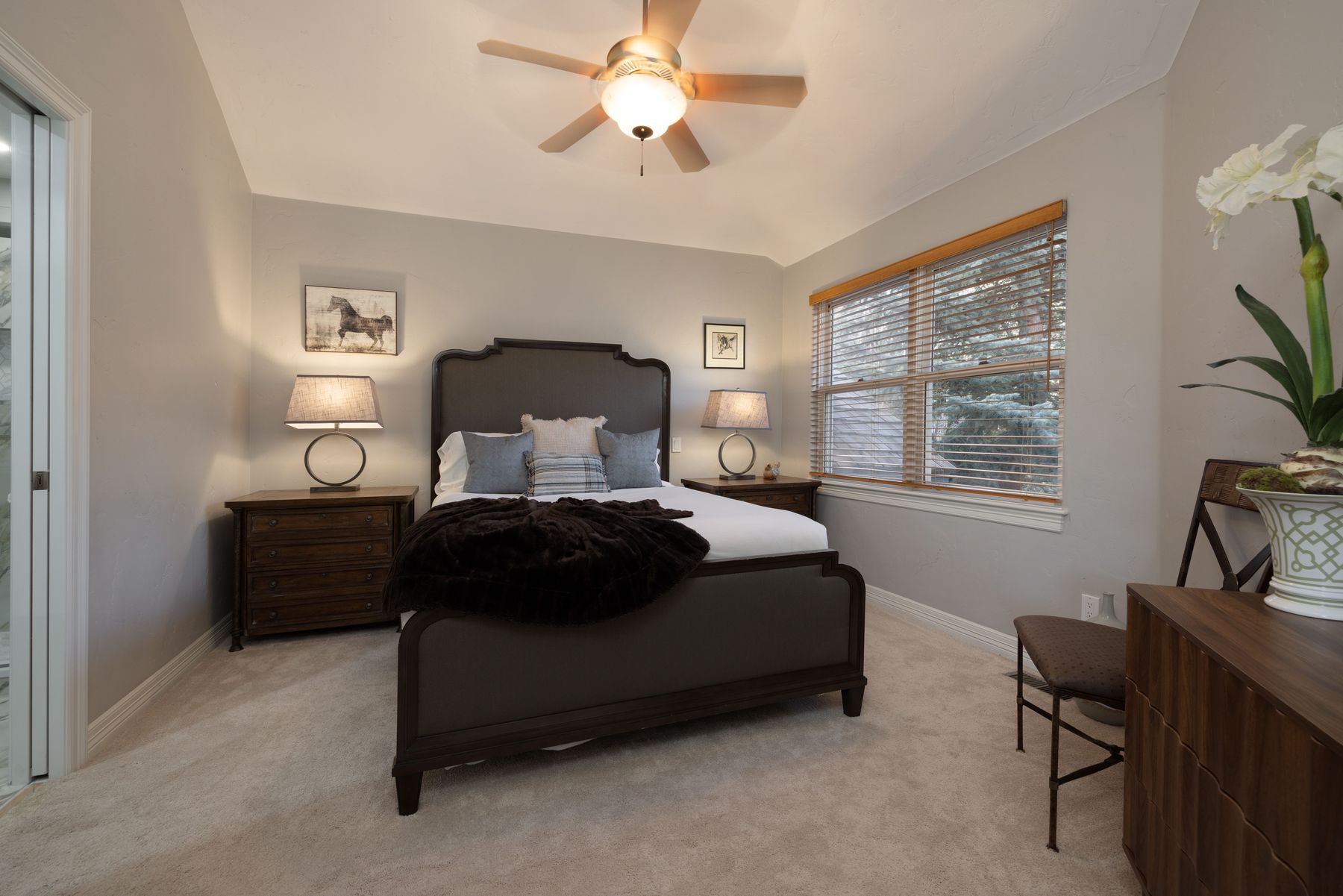 This cozy and well-appointed bedroom features a dark wood bed frame with a plush white mattress and a dark brown throw blanket. The room is accented by matching wooden nightstands with elegant lamps, a ceiling fan, and a window with wooden blinds that lets in natural light. A small vanity area with a chair and a decorative plant completes the inviting and neutral-toned space.