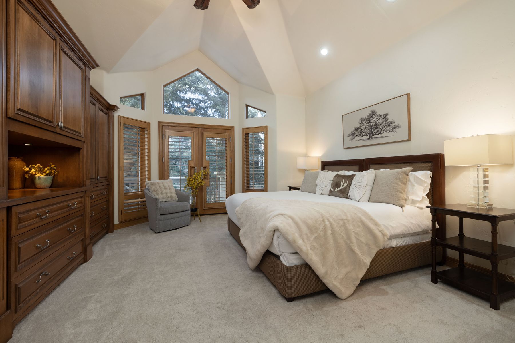 This spacious primary bedroom features a high vaulted ceiling with a large, multi-pane window that floods the room with natural light. The space is anchored by a large bed with a plush, cream-colored throw blanket and a dark wood headboard, complemented by a matching built-in wardrobe unit and a cozy gray armchair. The neutral color palette and soft carpeting create a warm, inviting atmosphere, while the view through the French doors suggests a serene, wooded setting.