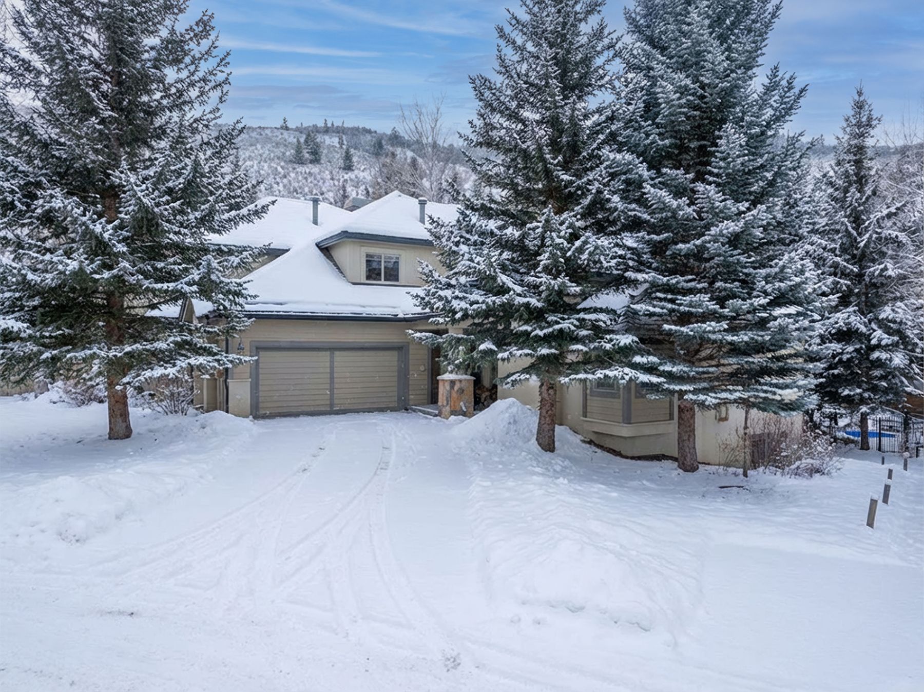 This front-facing exterior shot captures a charming, multi-level home nestled in a serene, snow-covered landscape. The property features a two-car garage, a welcoming entryway, and mature evergreen trees dusted with fresh snow, creating a cozy, winter-wonderland aesthetic. The perspective is eye-level, emphasizing the home's integration into its natural, mountainous surroundings.