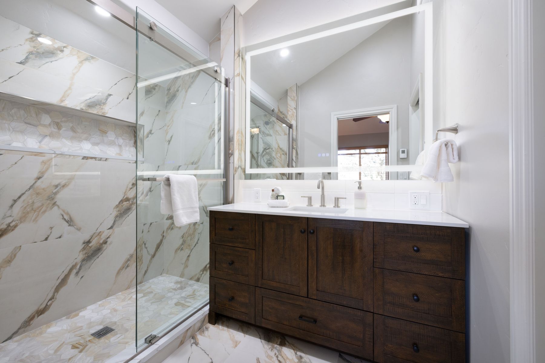 This elegant primary bathroom features a rustic dark wood vanity topped with a clean white countertop and a large, modern LED-backlit mirror. To the left, a spacious walk-in shower is encased in glass and adorned with luxurious marble-patterned tile and a hexagonal mosaic floor. The combination of warm wood tones and sophisticated stone finishes creates a high-end, spa-like atmosphere.