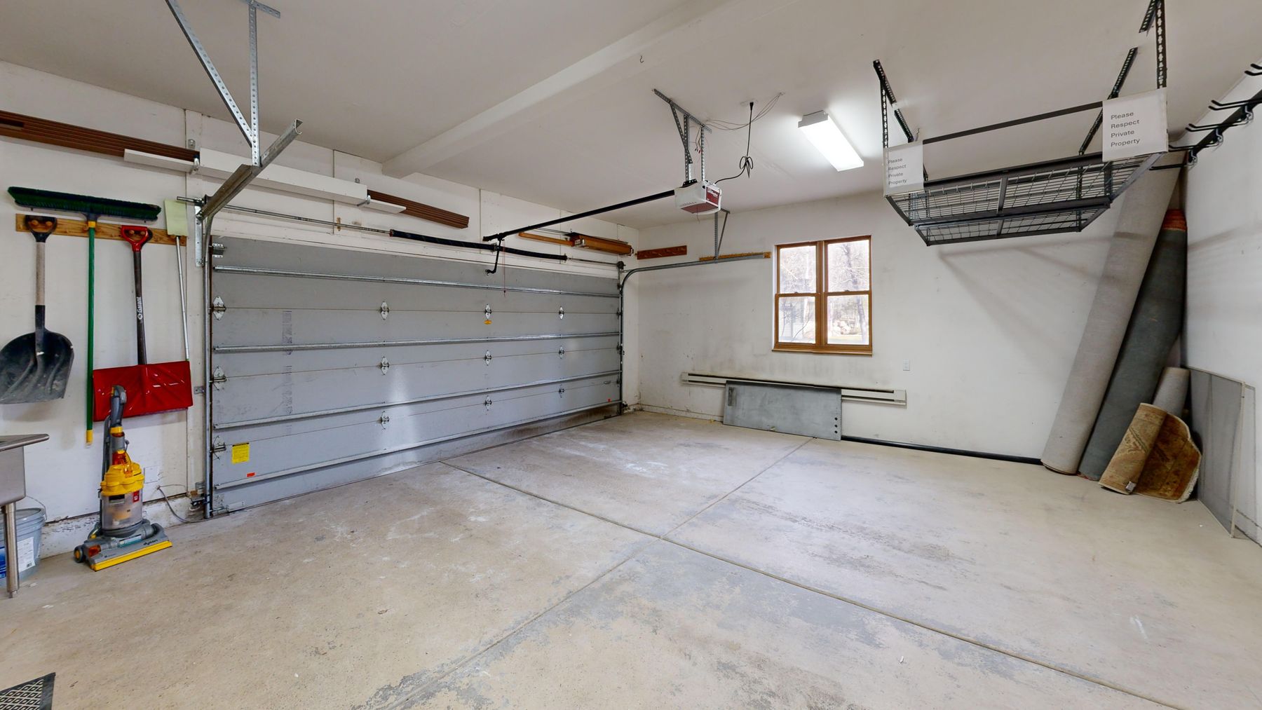 This image showcases a spacious, clean two-car garage with a large, light-gray sectional garage door and concrete flooring. The space includes practical storage solutions like a ceiling-mounted rack, wall-mounted tool organizers, and a window that provides natural light. The perspective is a wide-angle shot from the interior, emphasizing the depth and functional layout of the storage area.