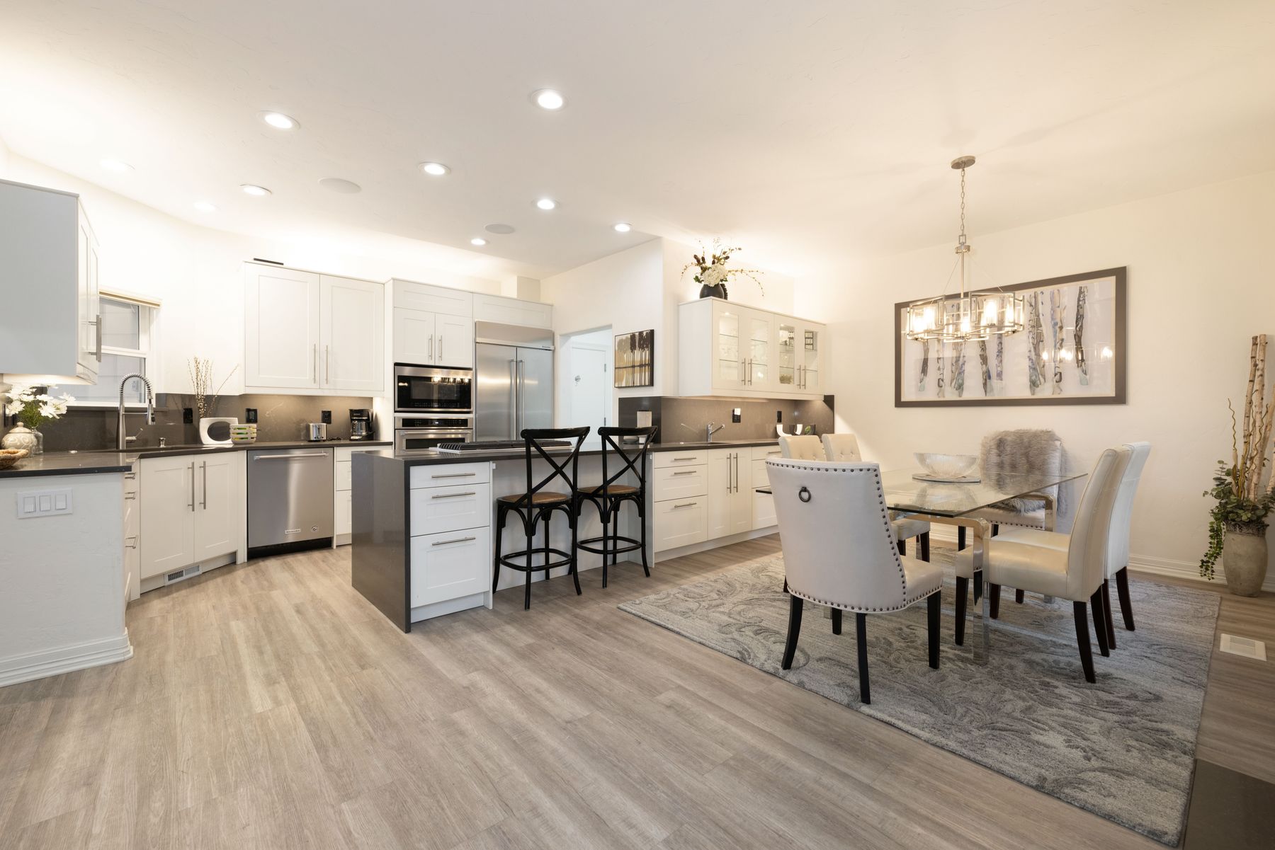 This bright and modern open-concept kitchen features crisp white cabinetry, sleek stainless steel appliances, and a contrasting dark gray kitchen island with bar seating. The space flows seamlessly into a dining area anchored by a glass-topped table and elegant upholstered chairs, all set against light wood-look flooring. The perspective is a wide-angle shot that captures the functional layout and sophisticated, airy aesthetic of the home.