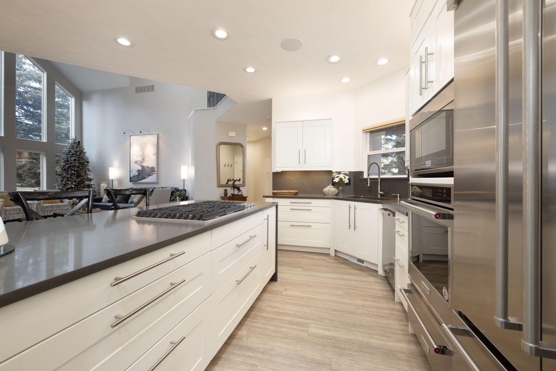 This modern, open-concept kitchen features sleek white cabinetry, dark quartz countertops, and high-end stainless steel appliances, including a built-in oven and microwave. The spacious island serves as a focal point, while the light wood-look flooring and recessed lighting create a bright, inviting atmosphere. The perspective looks from the kitchen island toward the adjacent living area, showcasing a seamless flow between spaces and a view of a large window with a holiday tree.