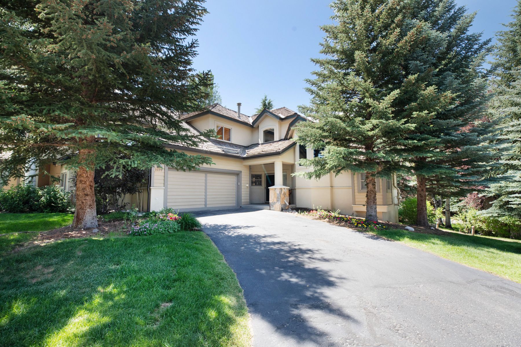 This inviting two-story home features a classic suburban design with a neutral-toned stucco facade and a prominent two-car garage. The property is framed by mature evergreen trees and a well-maintained lawn, creating a serene and established atmosphere. A paved driveway leads directly to the front entrance, which is accented by stone detailing, offering a warm and welcoming curb appeal.