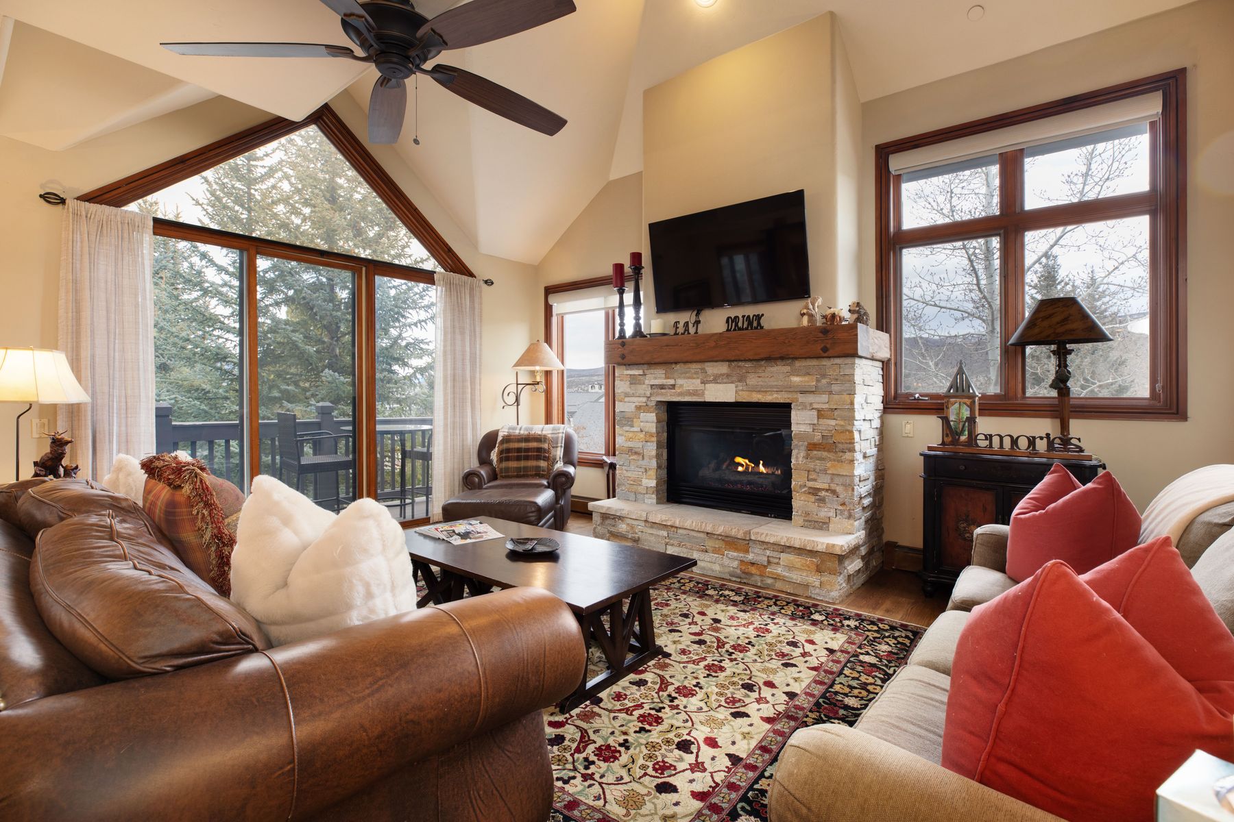 This cozy, rustic-style living room features a vaulted ceiling, a stone fireplace with a mounted television, and large windows that offer a scenic view of the surrounding trees. The space is furnished with a comfortable leather sofa, a plush beige sofa with red accent pillows, and a dark wood coffee table, all anchored by a traditional patterned area rug. The warm lighting and natural textures create an inviting, mountain-lodge atmosphere perfect for relaxation.