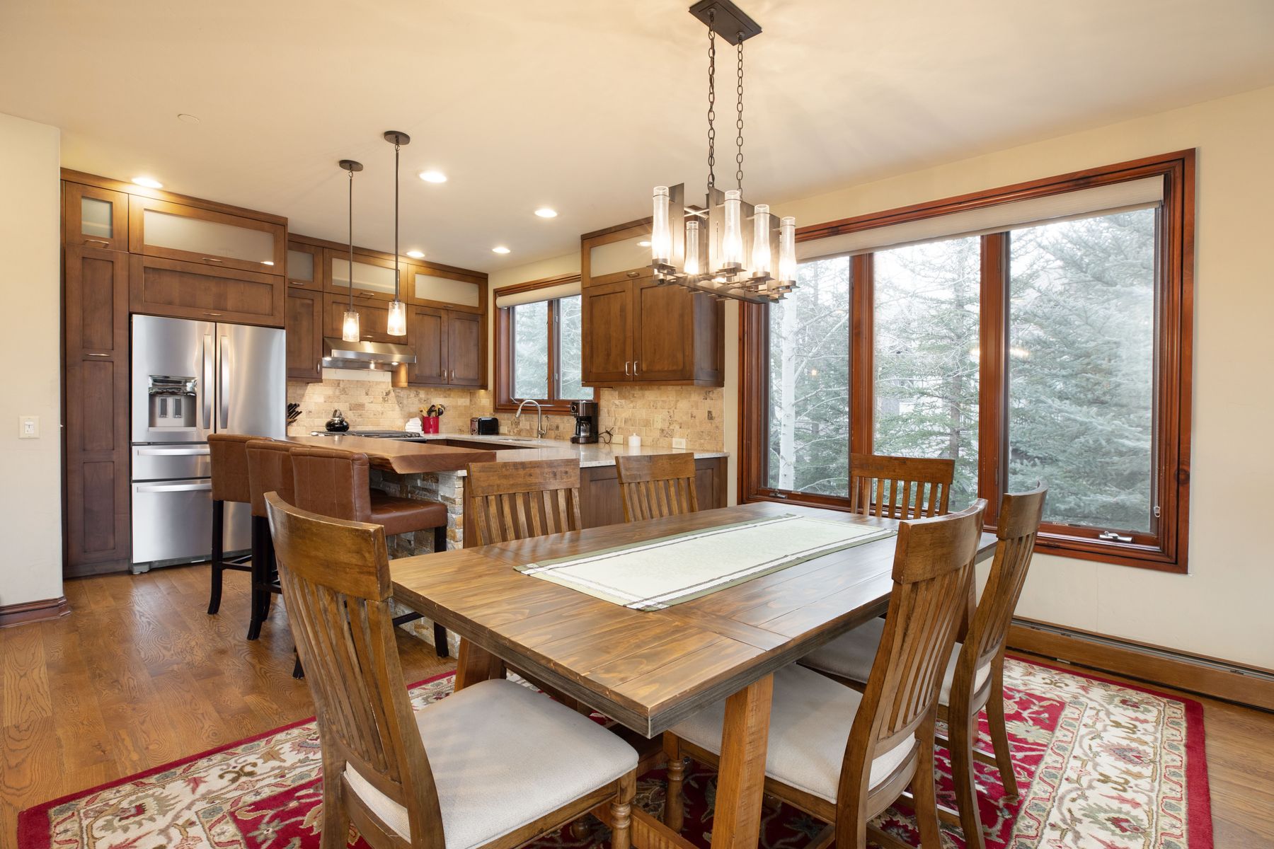 This open-concept space seamlessly blends a rustic dining area with a well-appointed kitchen, featuring warm wood cabinetry and a natural wood dining table. The room is anchored by a large, ornate chandelier and a patterned area rug, while expansive windows provide a scenic view of the surrounding forest. The overall aesthetic is cozy and inviting, emphasizing high-quality materials and a comfortable, mountain-lodge atmosphere.