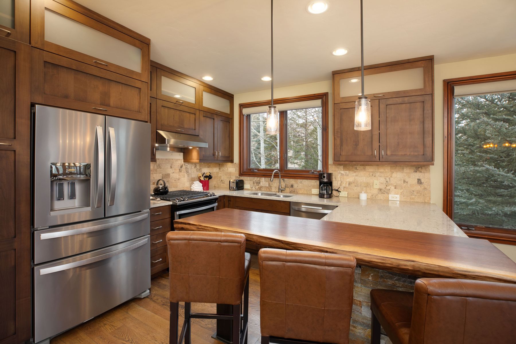 This warm and inviting kitchen features rich, dark wood cabinetry paired with a unique live-edge wood breakfast bar and stone-tiled backsplash. The space is equipped with modern stainless steel appliances, including a large refrigerator and a gas range, while pendant lights hang above the counter to create a cozy atmosphere. The perspective captures the functional layout, highlighting the seamless blend of rustic textures and contemporary convenience.