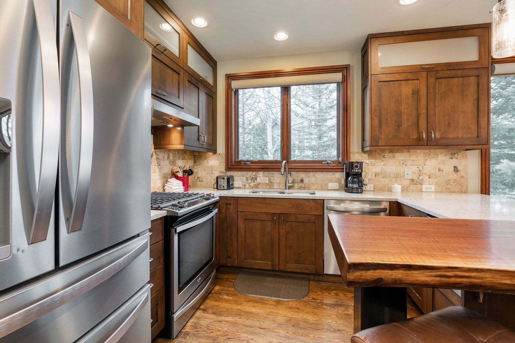 This warm and inviting kitchen features rich, dark wood cabinetry paired with light-colored granite countertops and a natural stone tile backsplash. The space is equipped with modern stainless steel appliances, including a large refrigerator and a gas range, while a live-edge wooden breakfast bar adds a rustic, custom touch. The perspective is a wide-angle shot from the kitchen entrance, highlighting the functional layout and the bright, natural light streaming in through the central window.