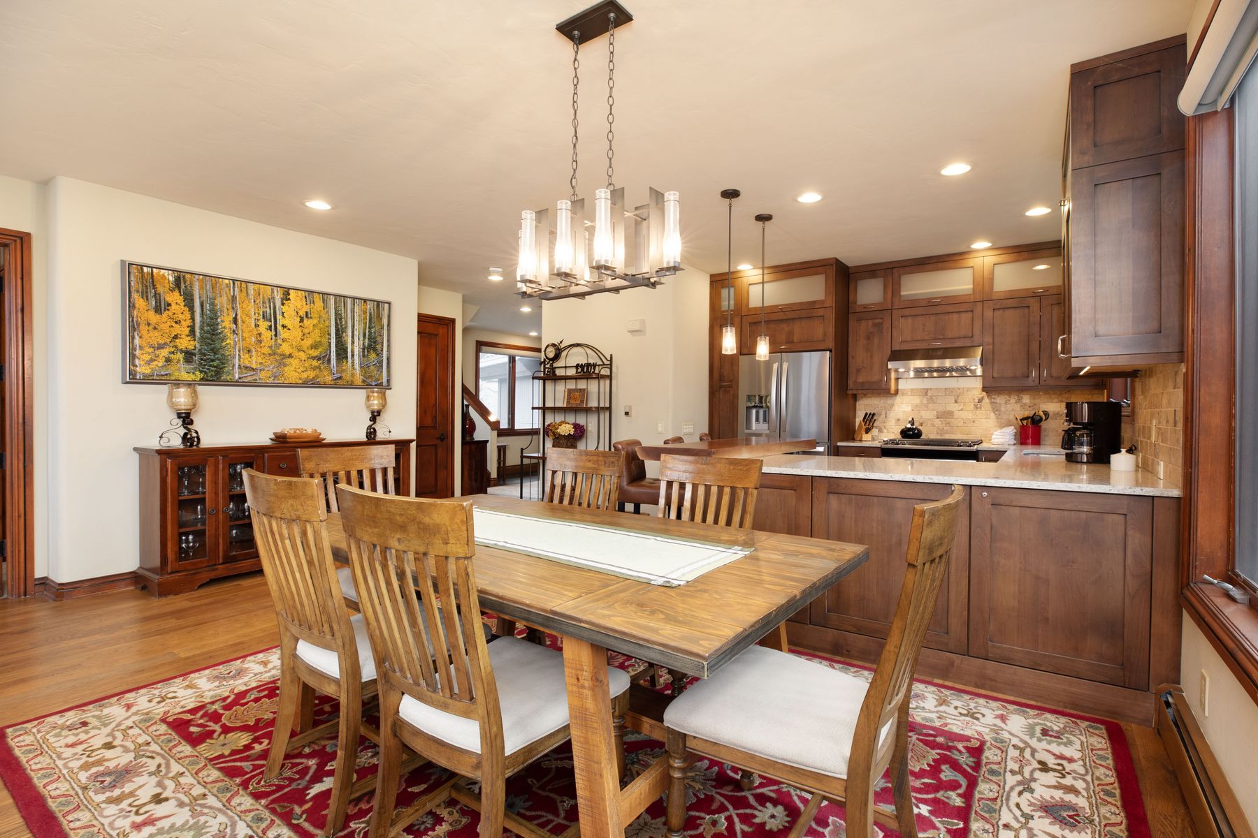 This open-concept dining area seamlessly transitions into a well-appointed kitchen, featuring warm wood cabinetry and granite countertops. A rustic wooden dining table serves as the centerpiece, complemented by a vibrant red patterned area rug and a modern linear chandelier. The space is bright and inviting, with a large landscape painting of an autumn forest adding a touch of natural color to the neutral walls.