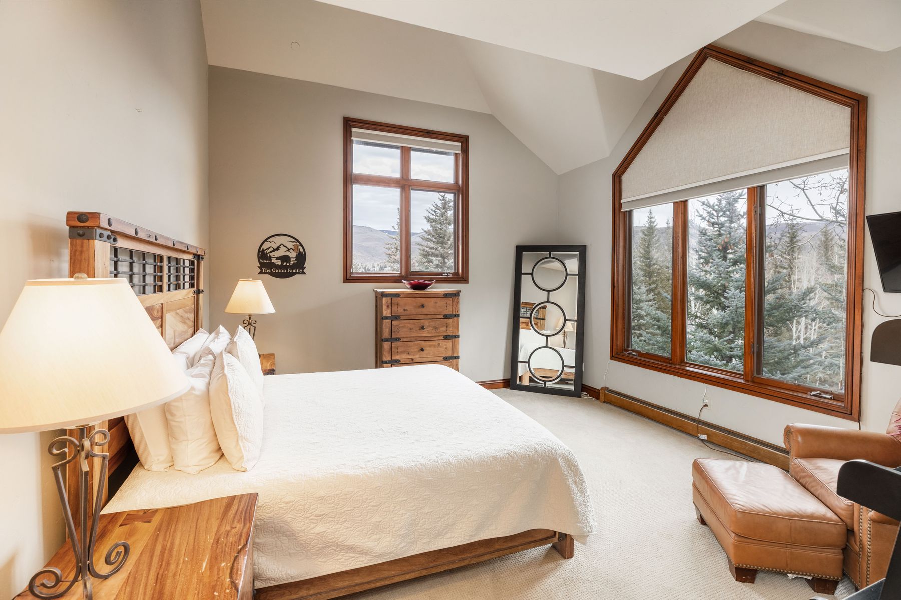 This spacious primary bedroom features a rustic-chic aesthetic with a wooden bed frame, matching nightstands, and a leather armchair. Large windows with wooden trim frame a scenic view of the surrounding forest, while a tall, circular-patterned floor mirror adds a modern touch to the room. The neutral color palette and vaulted ceilings create a serene and inviting atmosphere perfect for a mountain retreat.