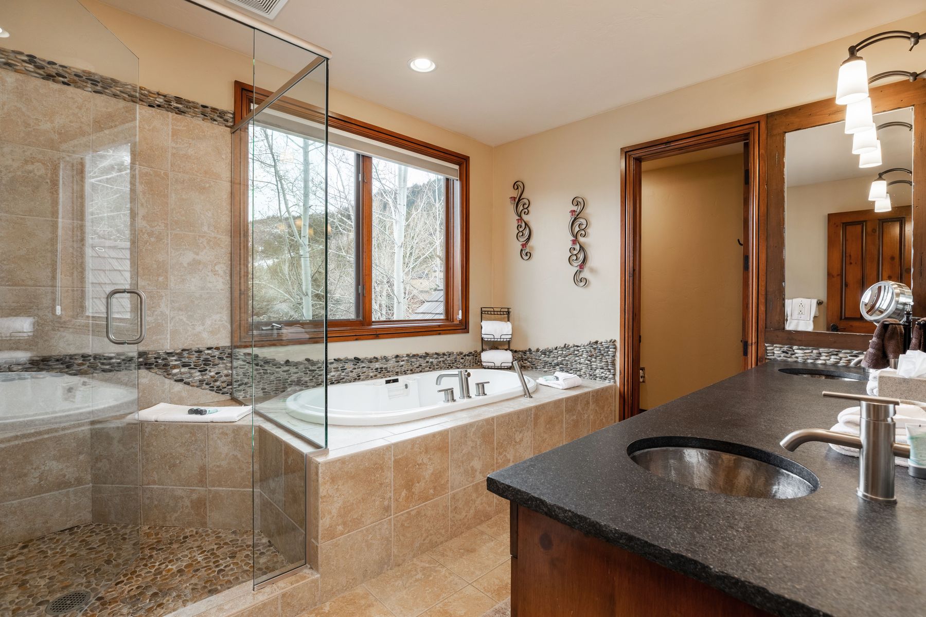 This spacious primary bathroom features a luxurious soaking tub nestled beneath a large window with scenic outdoor views, complemented by a separate glass-enclosed shower with pebble-stone flooring. The vanity area includes a dark granite countertop and warm wood cabinetry, creating a rustic yet elegant atmosphere. The room is accented with decorative wall art and soft recessed lighting, providing a serene and inviting spa-like environment.