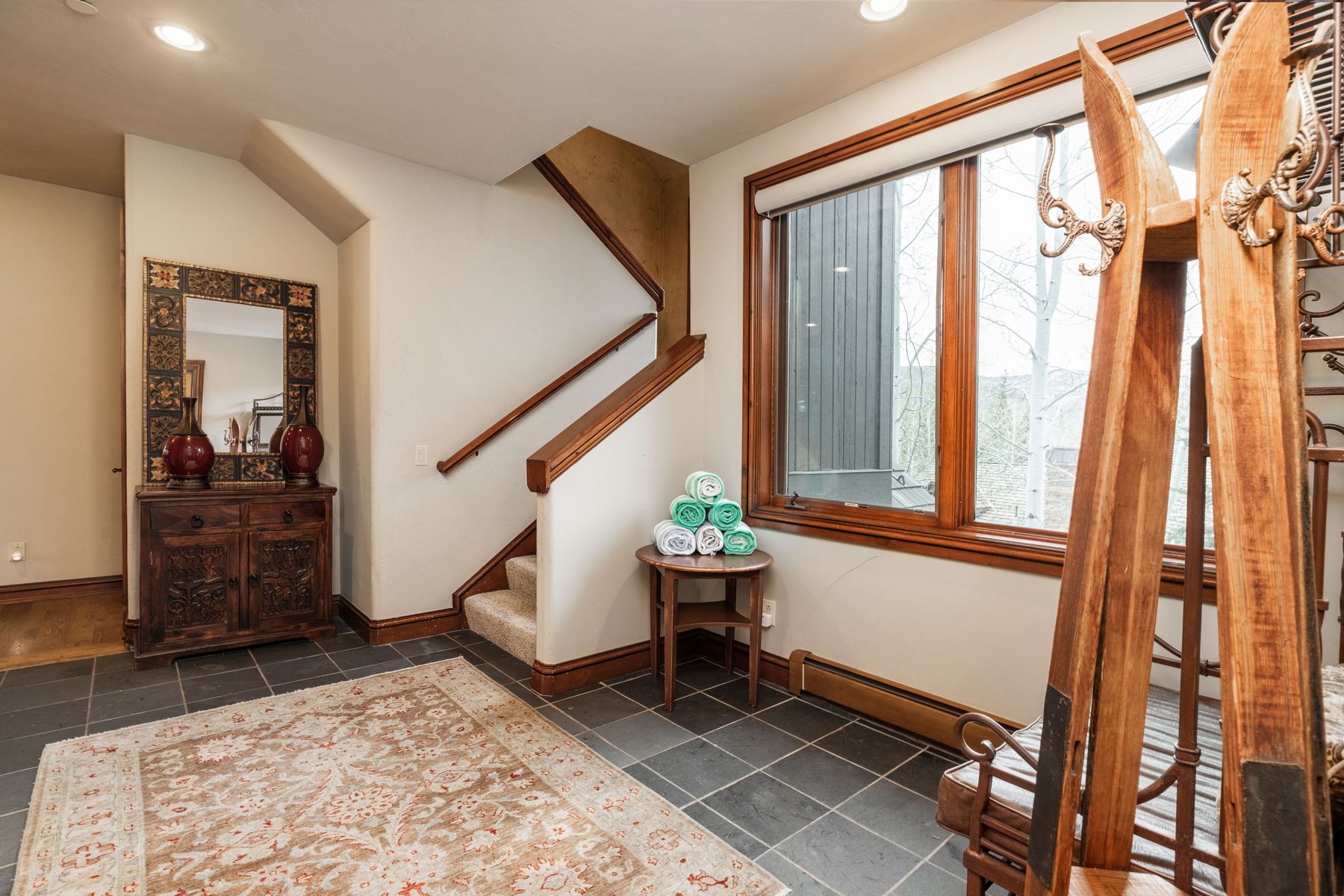 This inviting entryway features a rustic aesthetic with slate tile flooring, a decorative wooden cabinet topped with a mirror, and a staircase leading to the upper level. A small round table holds a stack of rolled towels, while a large window provides natural light and a view of the outdoors. The space is accented by a patterned area rug and a unique wooden coat rack, creating a warm and welcoming atmosphere.
