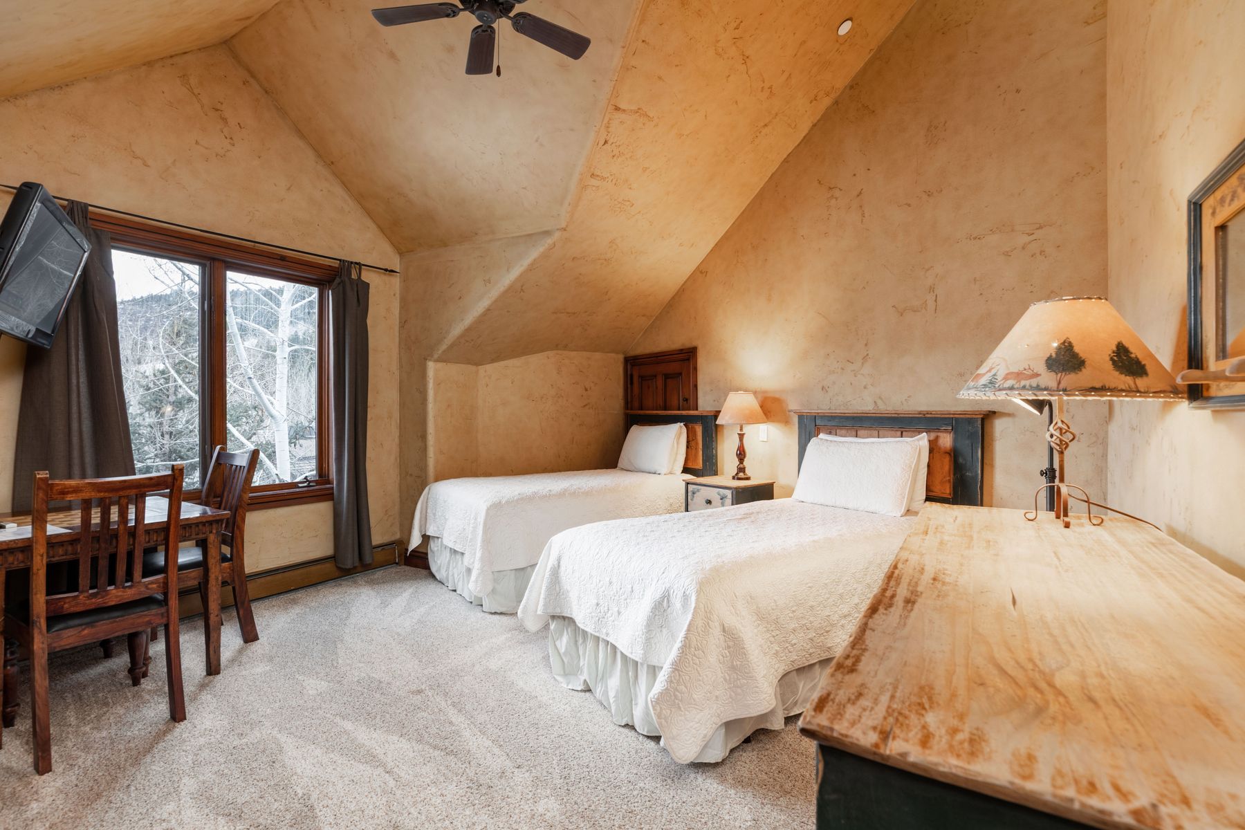 This cozy guest bedroom features a rustic, mountain-lodge aesthetic with textured, warm-toned walls and a vaulted ceiling. The room is furnished with two twin beds, a wooden desk with a chair, and a decorative lamp, all set against a backdrop of a snowy outdoor view through the window. The overall atmosphere is inviting and comfortable, perfect for a mountain retreat.