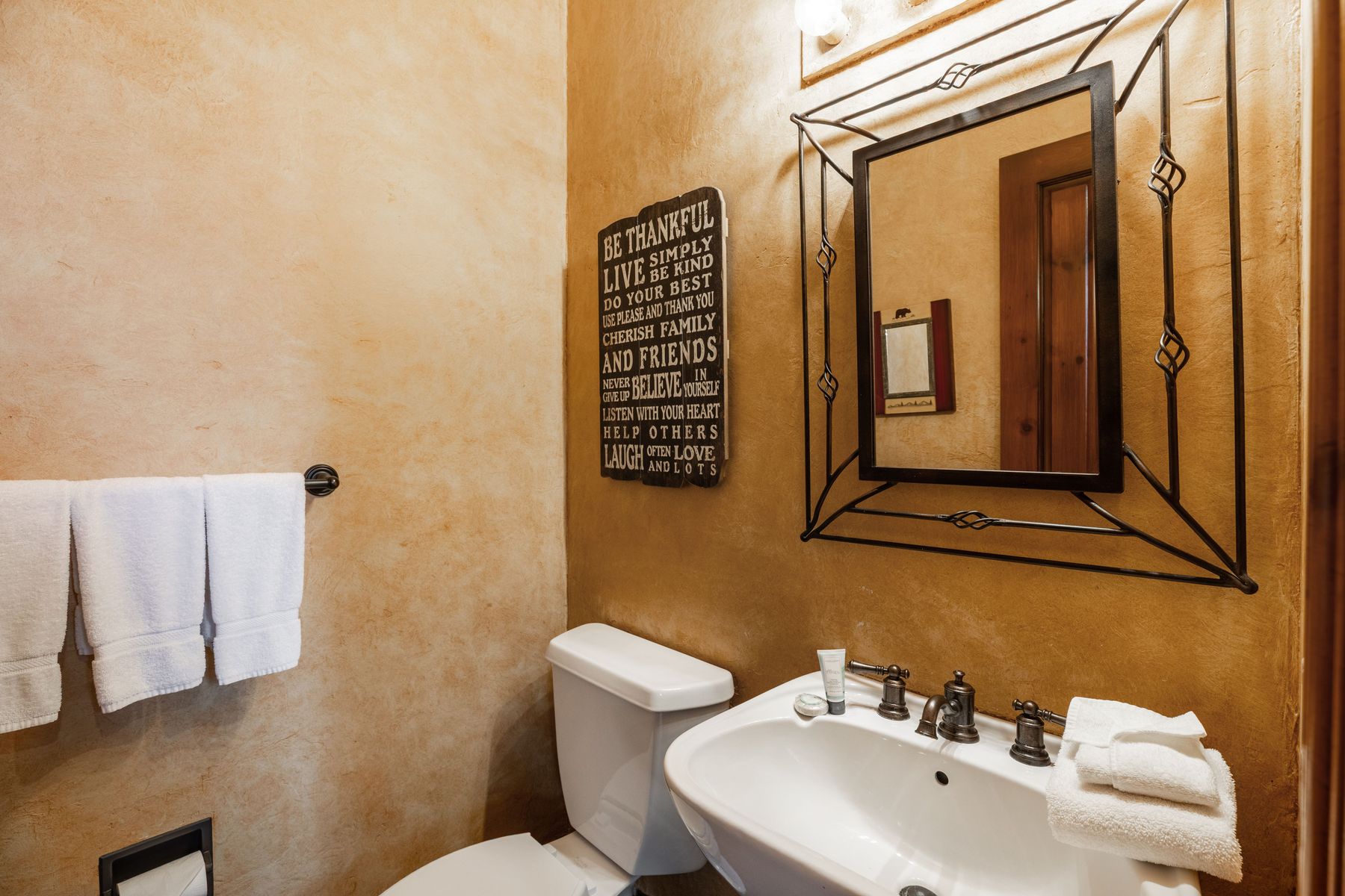 This cozy powder room features warm, textured faux-finish walls and a rustic wrought-iron framed mirror that adds a touch of mountain-lodge charm. A white pedestal sink with bronze fixtures sits beneath a decorative wall sign, while plush white towels provide a clean, inviting contrast. The space is well-lit and thoughtfully decorated, creating a welcoming atmosphere for guests.