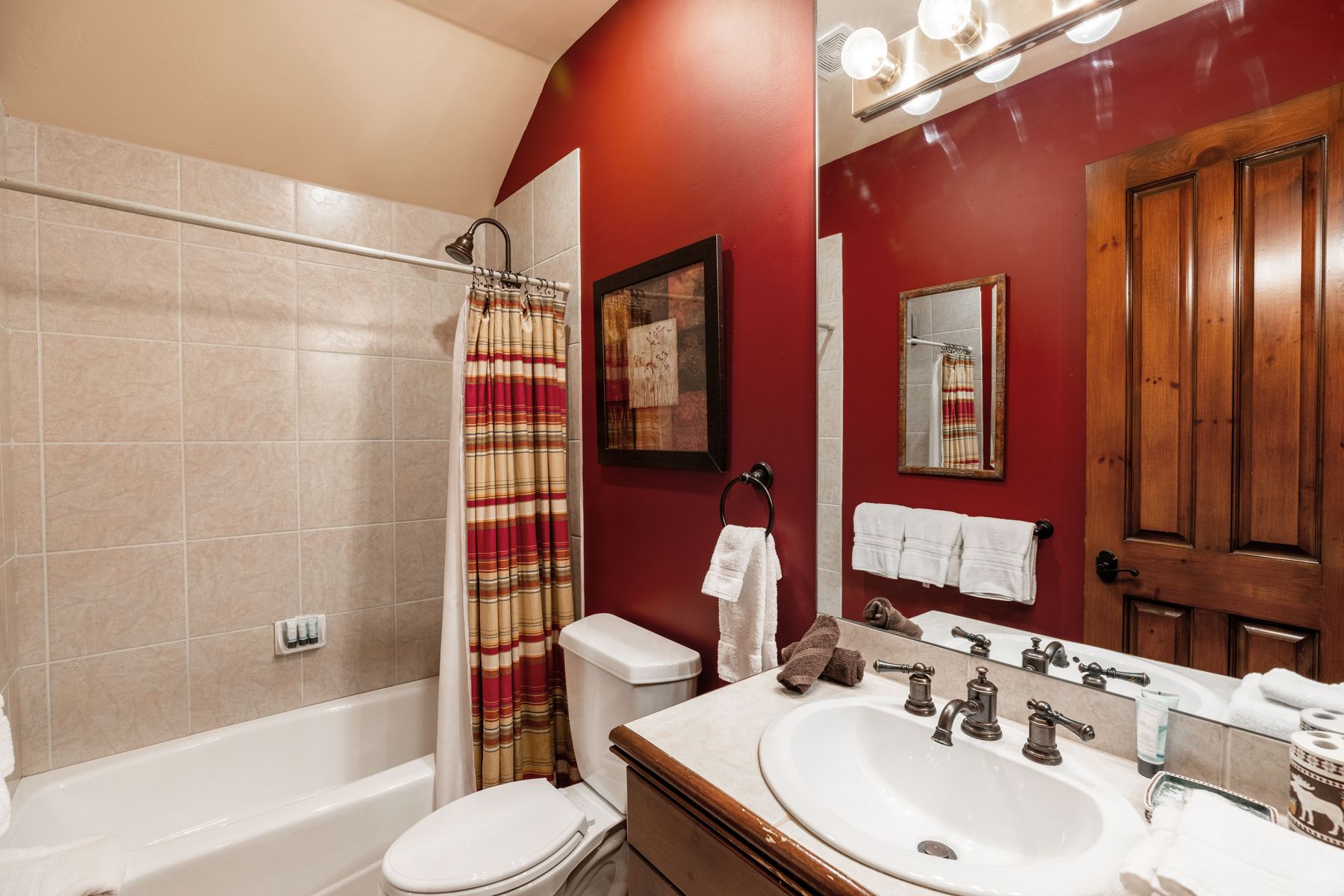 This bathroom features a bold red accent wall paired with neutral beige tiling in the shower-tub combination. The space includes a wooden vanity with a white sink, a large mirror, and a classic shower curtain, creating a warm and rustic aesthetic. The perspective is a wide-angle shot from the doorway, capturing the functional layout and cozy atmosphere of the room.