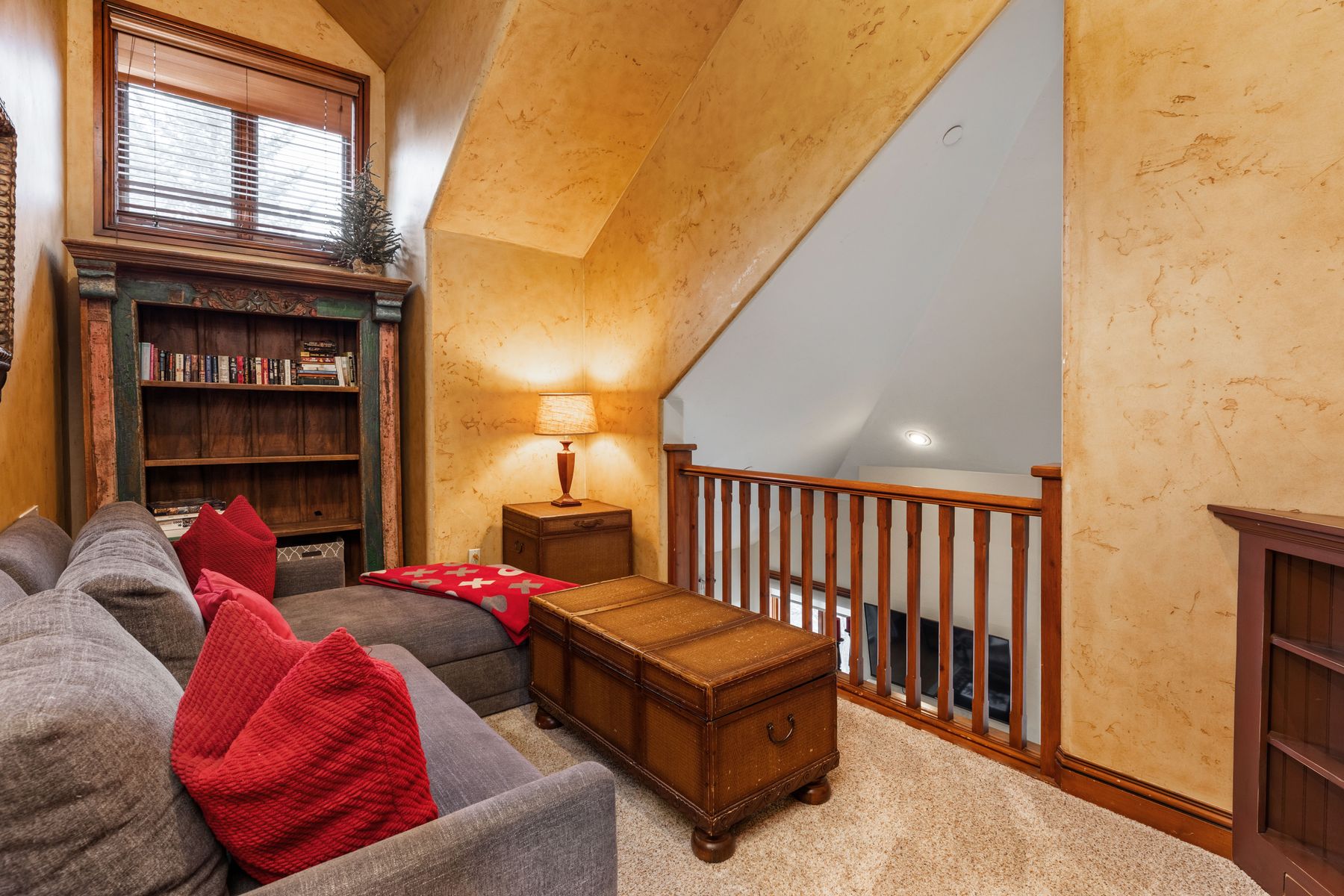 This cozy attic loft space features a comfortable gray sectional sofa adorned with vibrant red pillows, positioned next to a rustic wooden trunk coffee table. The room is accented by warm, textured yellow walls, a built-in bookshelf, and a wooden railing that overlooks the lower level of the home. The slanted ceiling and natural light from the window create an inviting, intimate reading nook or relaxation area.