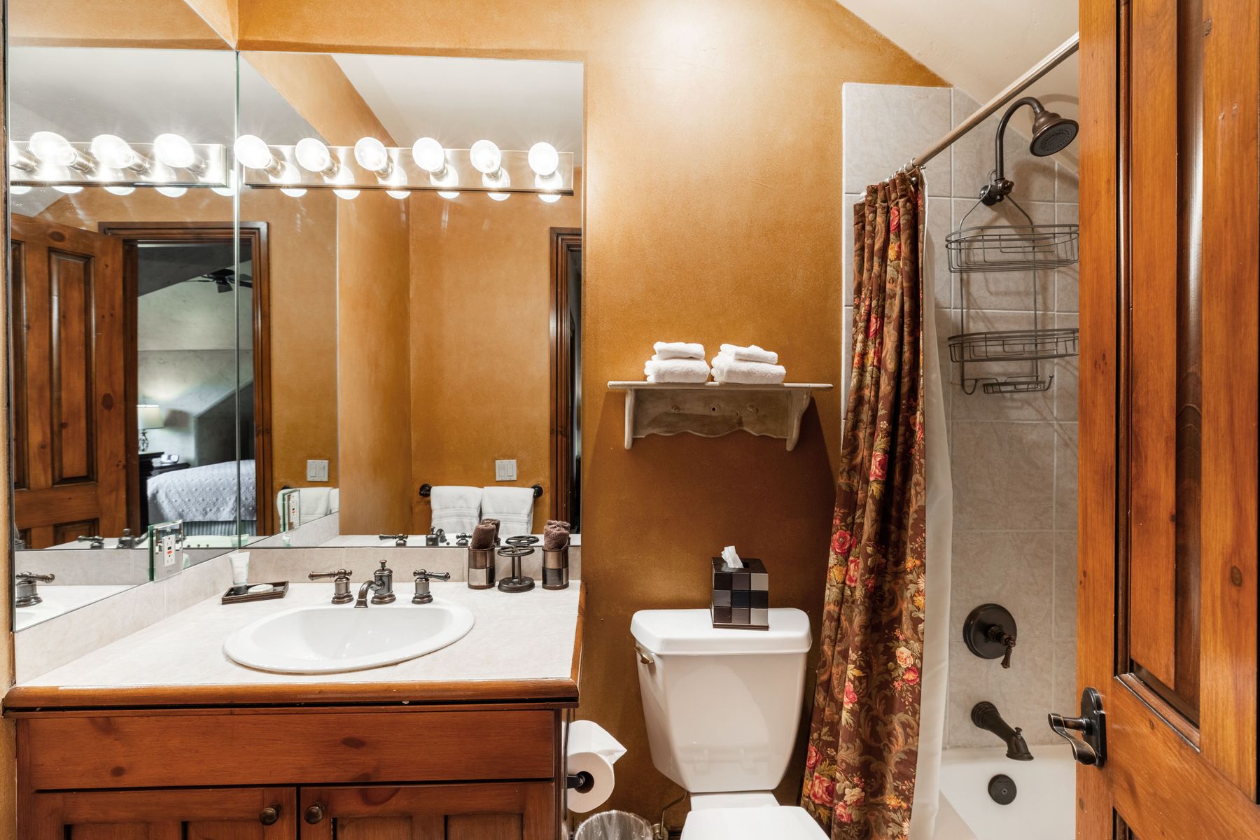 This cozy bathroom features a warm, golden-toned wall finish paired with a wooden vanity and a white porcelain sink. A large mirror spans the wall above the vanity, reflecting the adjacent bedroom, while a floral shower curtain adds a touch of traditional charm to the tub-shower combination. The space is functional and well-lit, offering a comfortable and inviting atmosphere.
