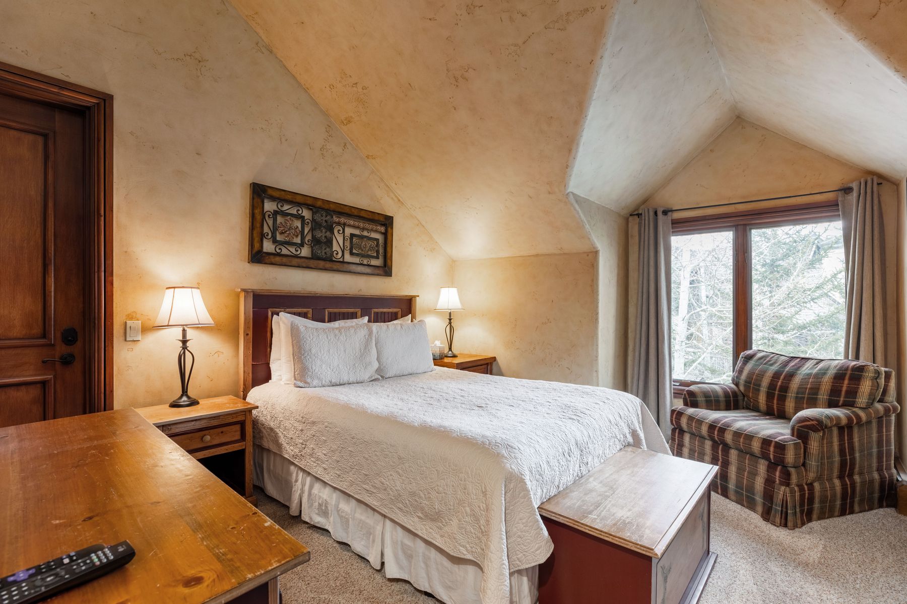 This cozy bedroom features a rustic, warm aesthetic with textured, earth-toned walls and a vaulted ceiling. The room is furnished with a queen-sized bed, a plaid armchair, and a wooden storage chest at the foot of the bed, all illuminated by soft bedside lamps. A large window with neutral curtains provides a view of the outdoors, creating a peaceful and inviting retreat.