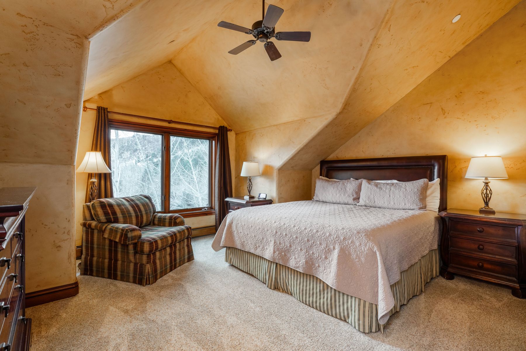 This cozy primary bedroom features a vaulted ceiling with a rustic, textured finish and a central ceiling fan. The room is anchored by a large bed with a dark leather headboard, flanked by matching wooden nightstands and lamps, while a plaid armchair sits near the window. The warm, earthy color palette and soft carpeting create a comfortable and inviting atmosphere.