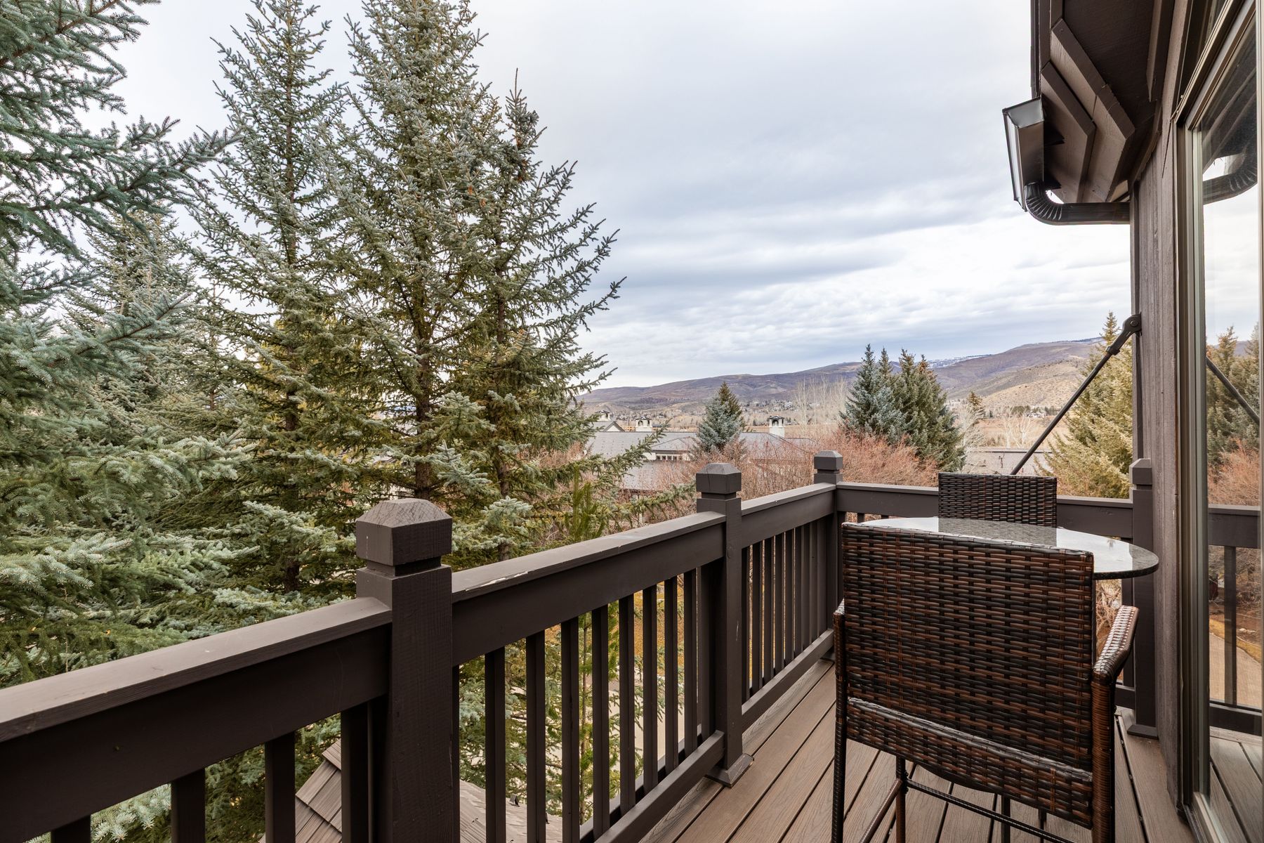 This elevated balcony offers a serene, scenic view of a mountainous landscape and dense evergreen trees. The space is furnished with a small, modern wicker bistro set, perfect for enjoying the quiet outdoor atmosphere. The dark wood railing and decking provide a rustic, cohesive aesthetic that complements the natural surroundings.