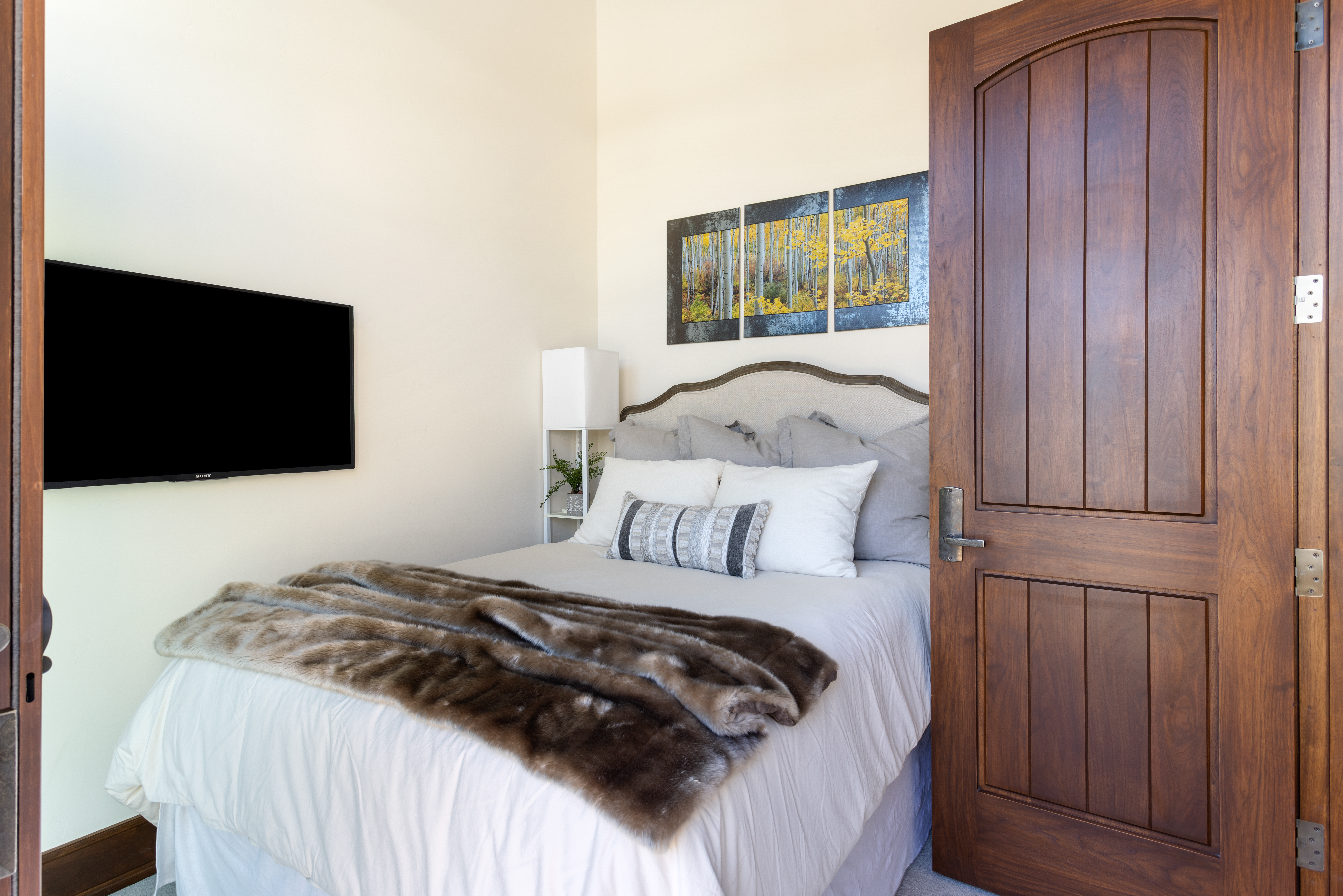This is a cozy bedroom featuring a queen-sized bed with a neutral-toned headboard and bedding, complemented by decorative pillows and a faux fur throw. A flat-screen TV is mounted on the wall, and artwork hangs above the bed. The room is well-lit and has a warm, inviting atmosphere, enhanced by a dark wood door.