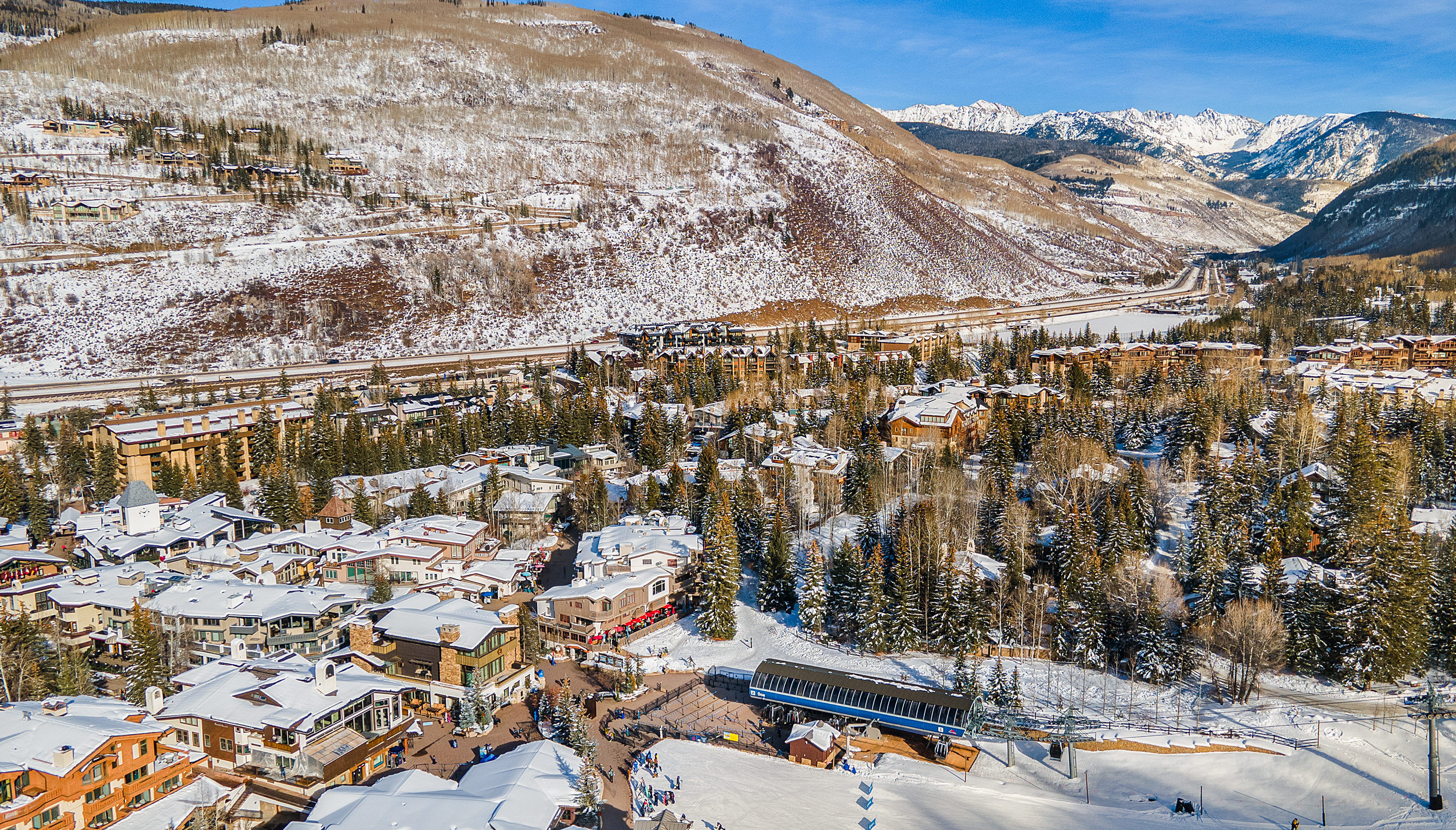 This aerial view showcases a luxurious ski resort town nestled in a snowy mountain valley. The scene features a mix of residential buildings and hotels with snow-covered roofs, surrounded by evergreen trees and ski slopes. In the background, majestic snow-capped mountains rise against a clear blue sky, creating a picturesque and inviting atmosphere for potential buyers seeking a winter retreat.